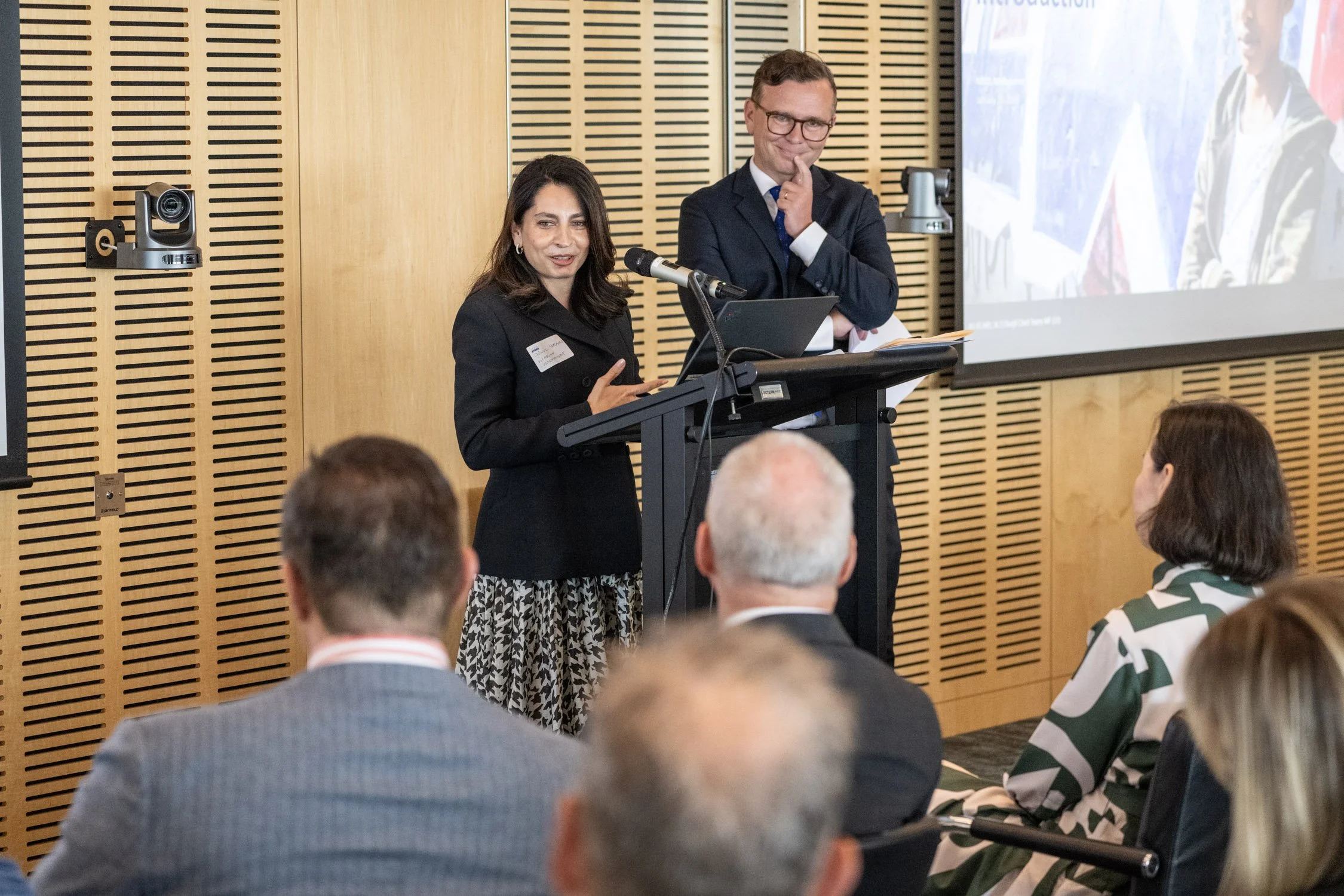 A woman speaking at a podium during a conference, with a man standing behind her. Both are dressed professionally and addressing an audience. The setting is a modern conference room with wooden paneling and a projector screen displaying a presentation.