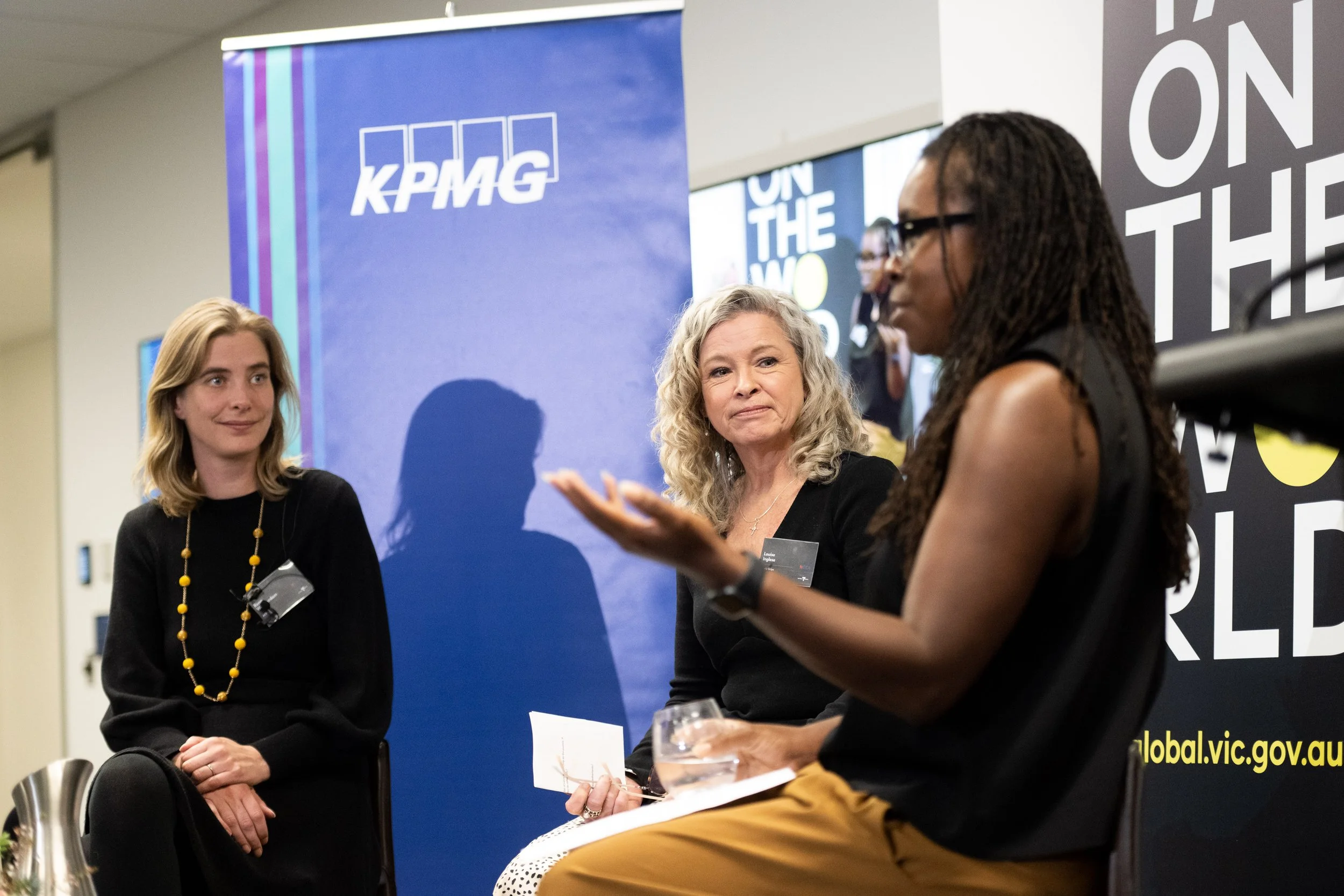 Three women seated and engaging in a discussion at an event with a KPMG banner in the background.