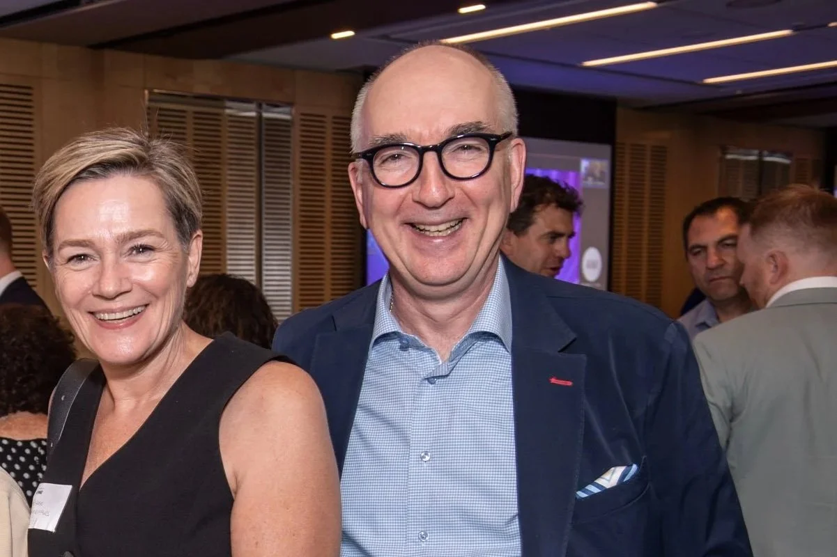 Smiling middle-aged woman and man attending a social event or conference indoors, people mingling in the background.