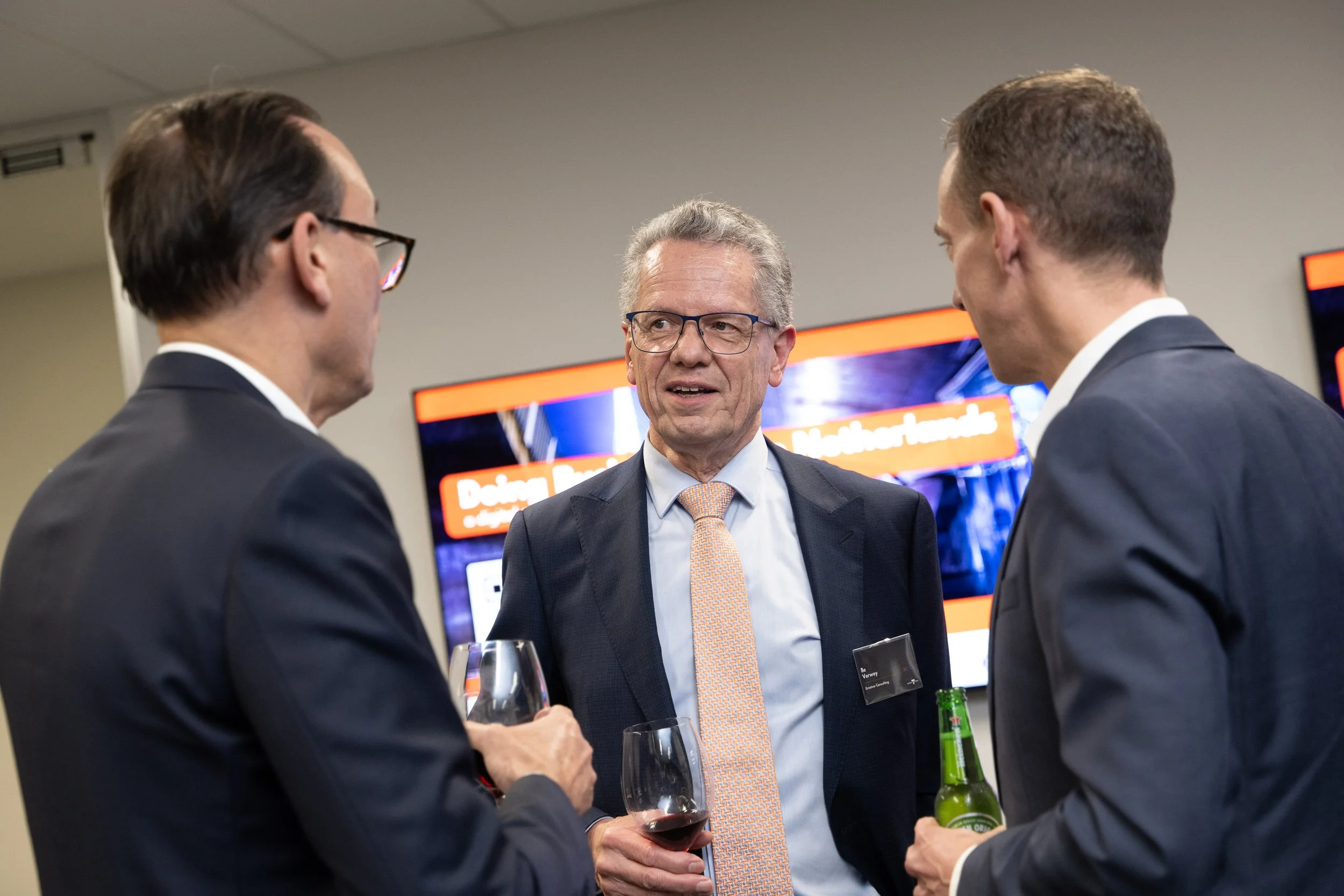 Three men in suits and glasses engaged in conversation at a formal event, holding drinks, with a large screen displaying colorful graphics behind them.