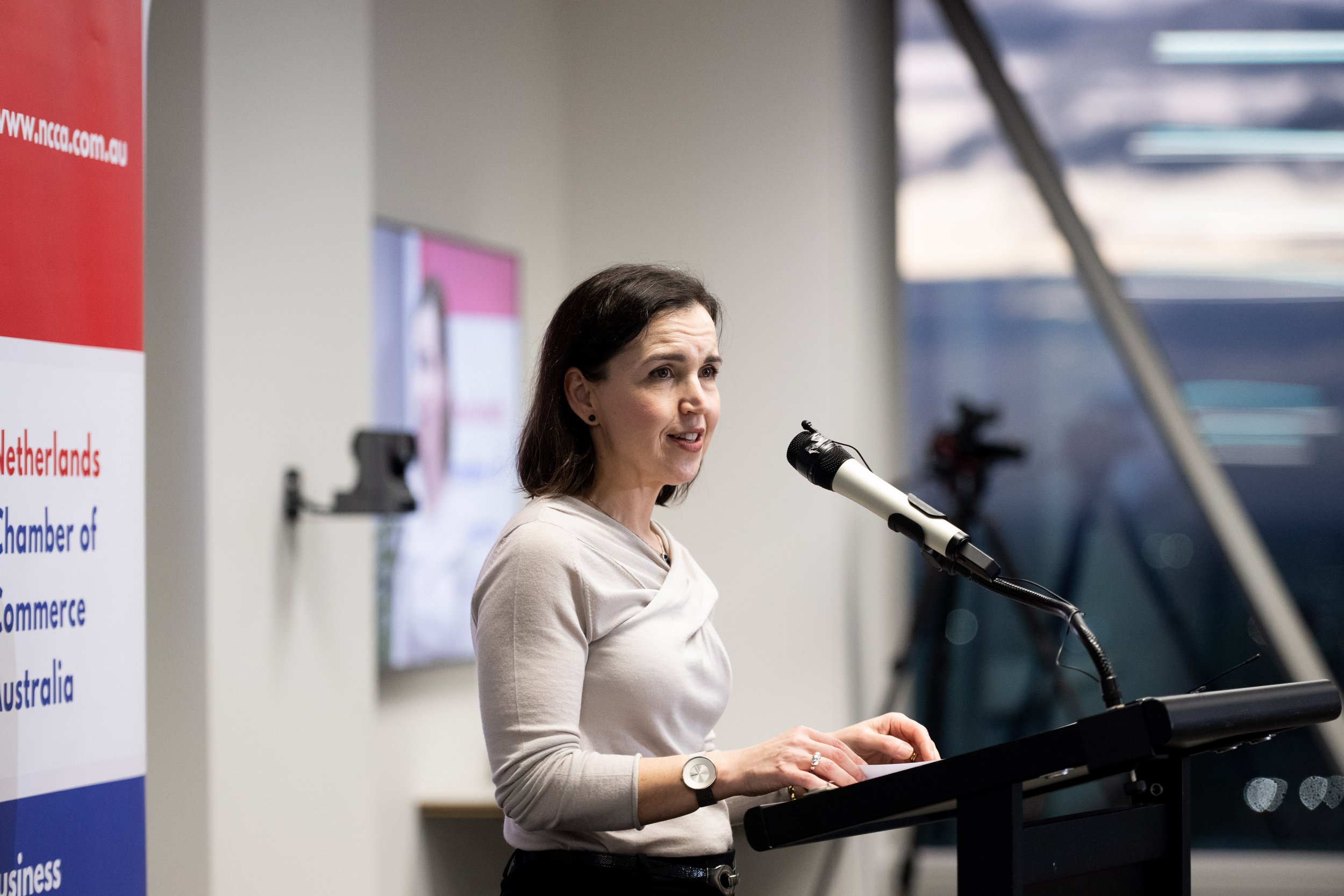 A woman stands at a lectern giving a speech, with a microphone in front of her. She has shoulder-length dark hair and is wearing a light-colored top.
