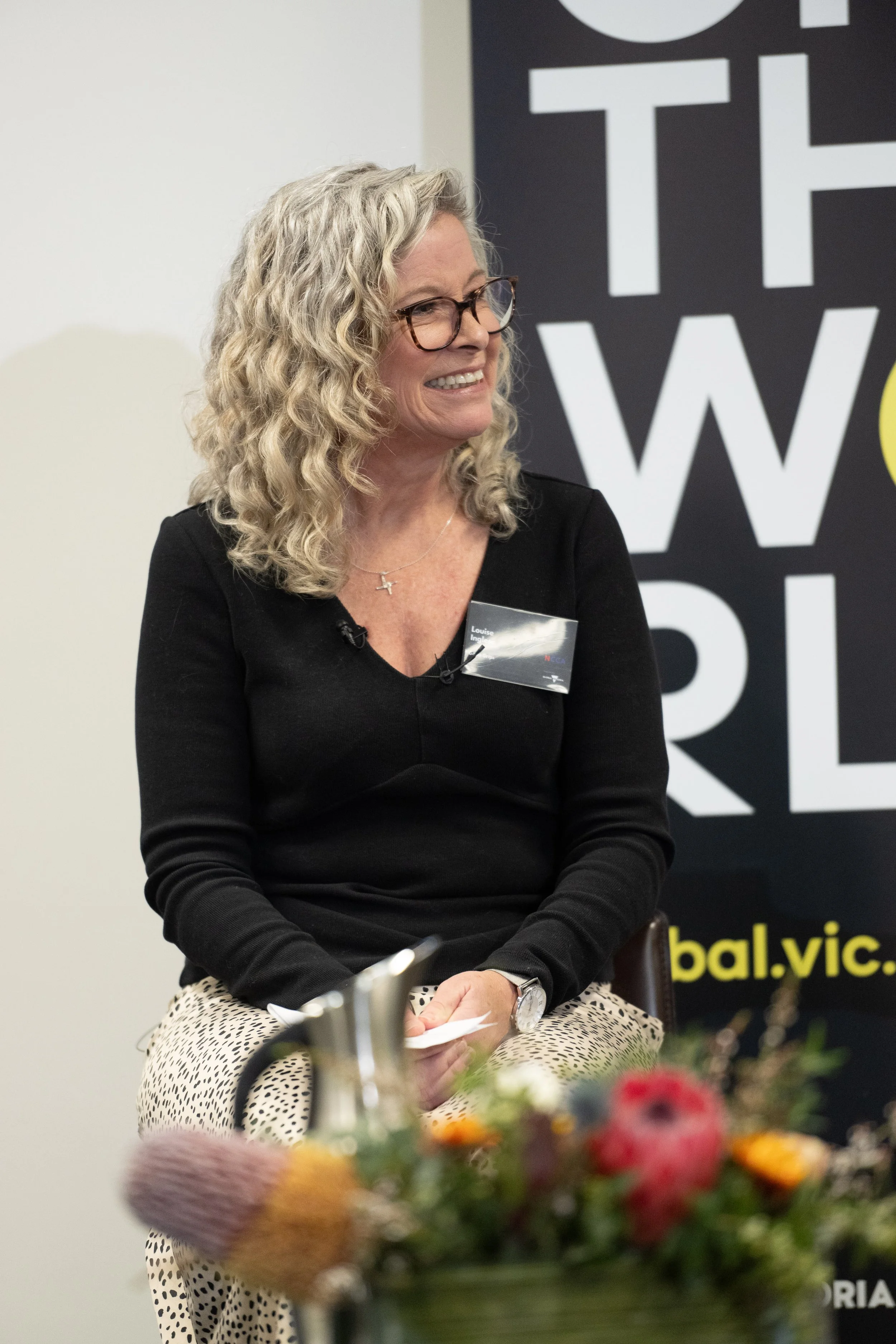 A woman with curly blonde hair, glasses, and a black top, smiling during an event with a banner in the background that reads "The World" and a flower arrangement in the foreground.