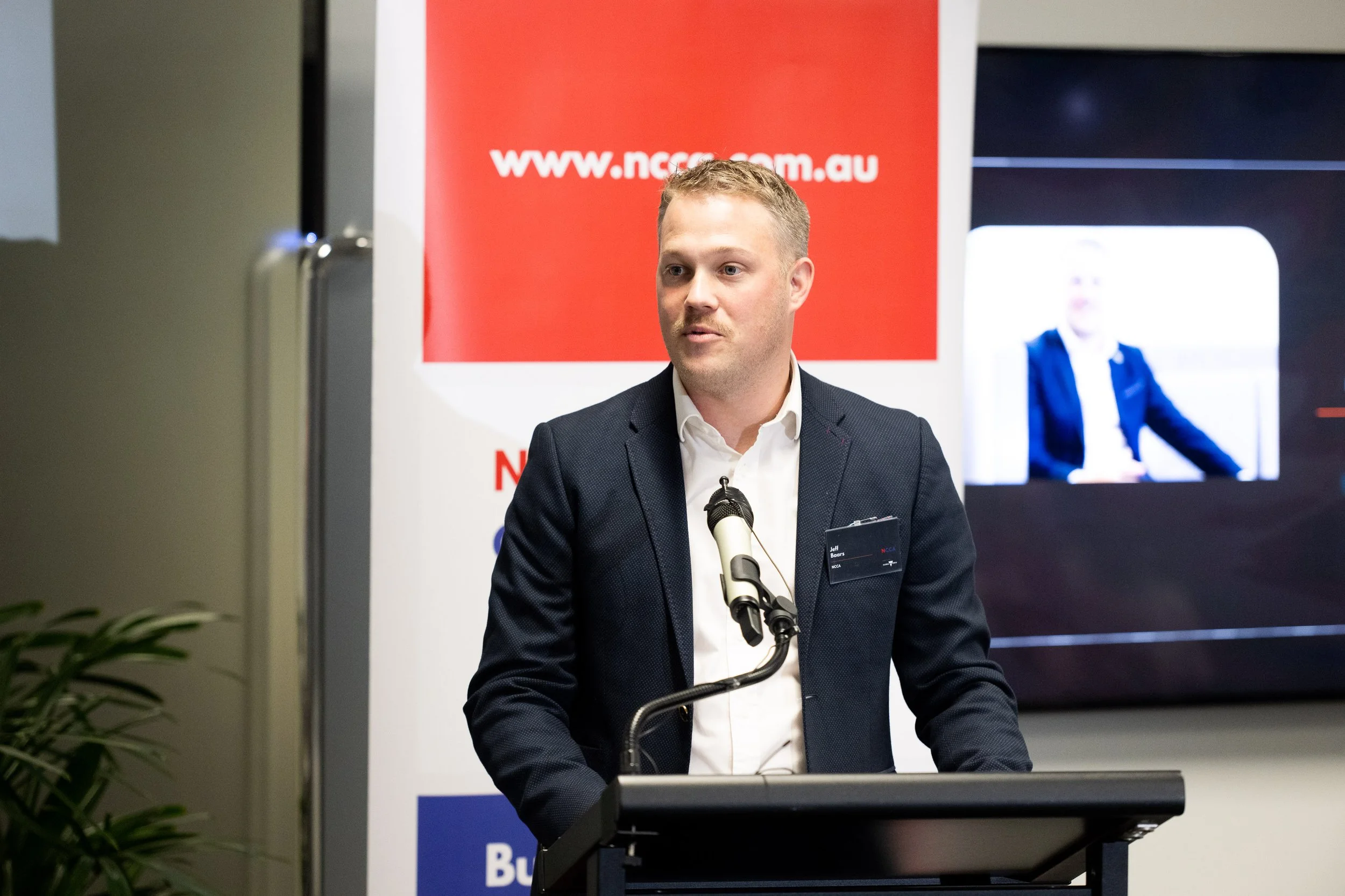 A man wearing a dark blazer and white shirt speaking at a podium during a presentation or event. There is a screen behind him showing his image and a red sign with a website URL.