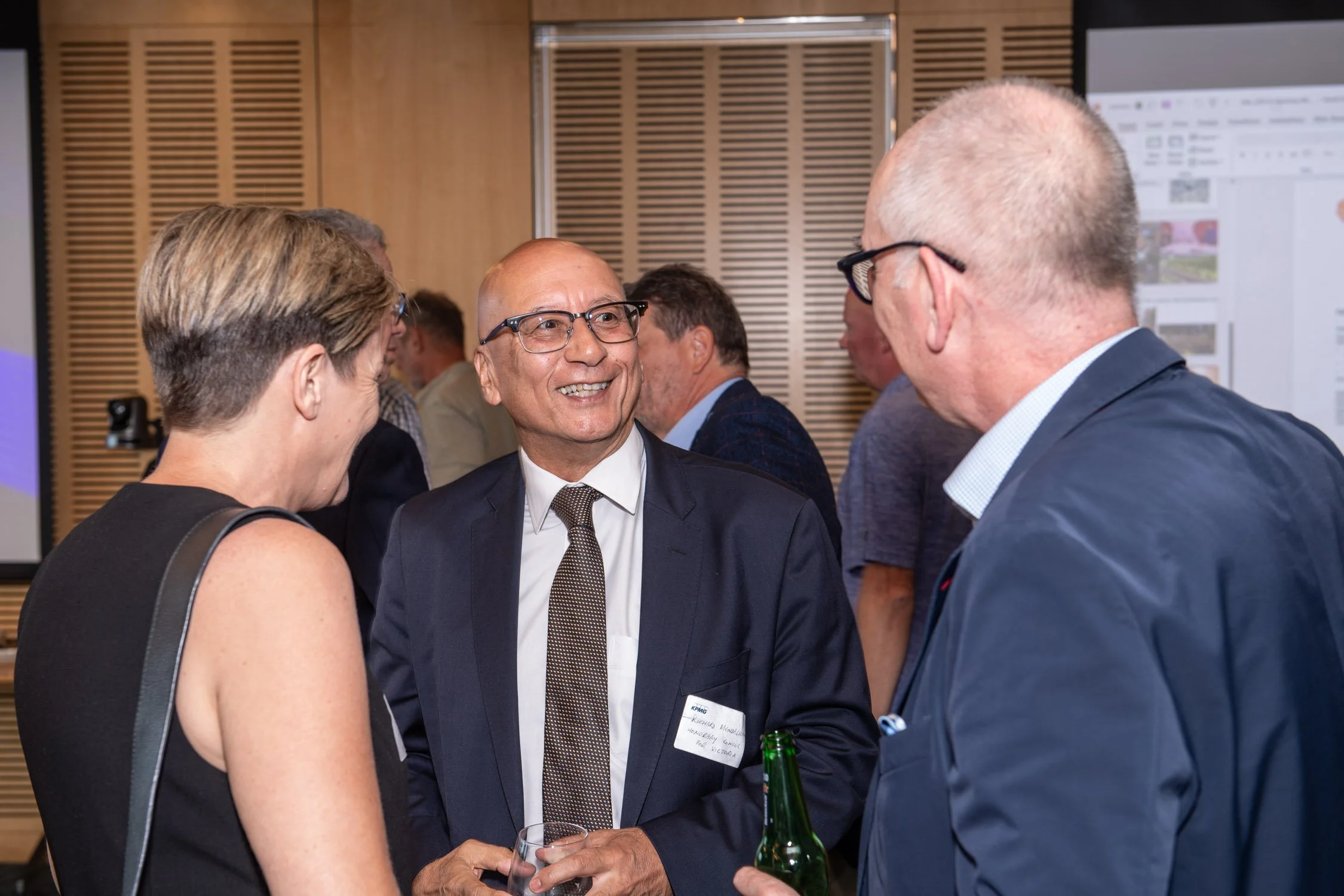 Three people engaged in conversation at a professional event, with a woman on the left and two men facing her, one holding a drink and the other a beverage bottle, in a room with wood-paneled walls and presentation posters.