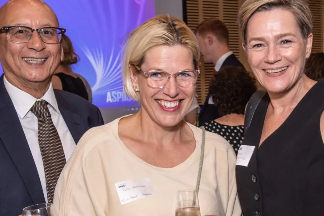 Three smiling adults at a formal event with a blue and purple backdrop, wearing business attire, and holding drinks.