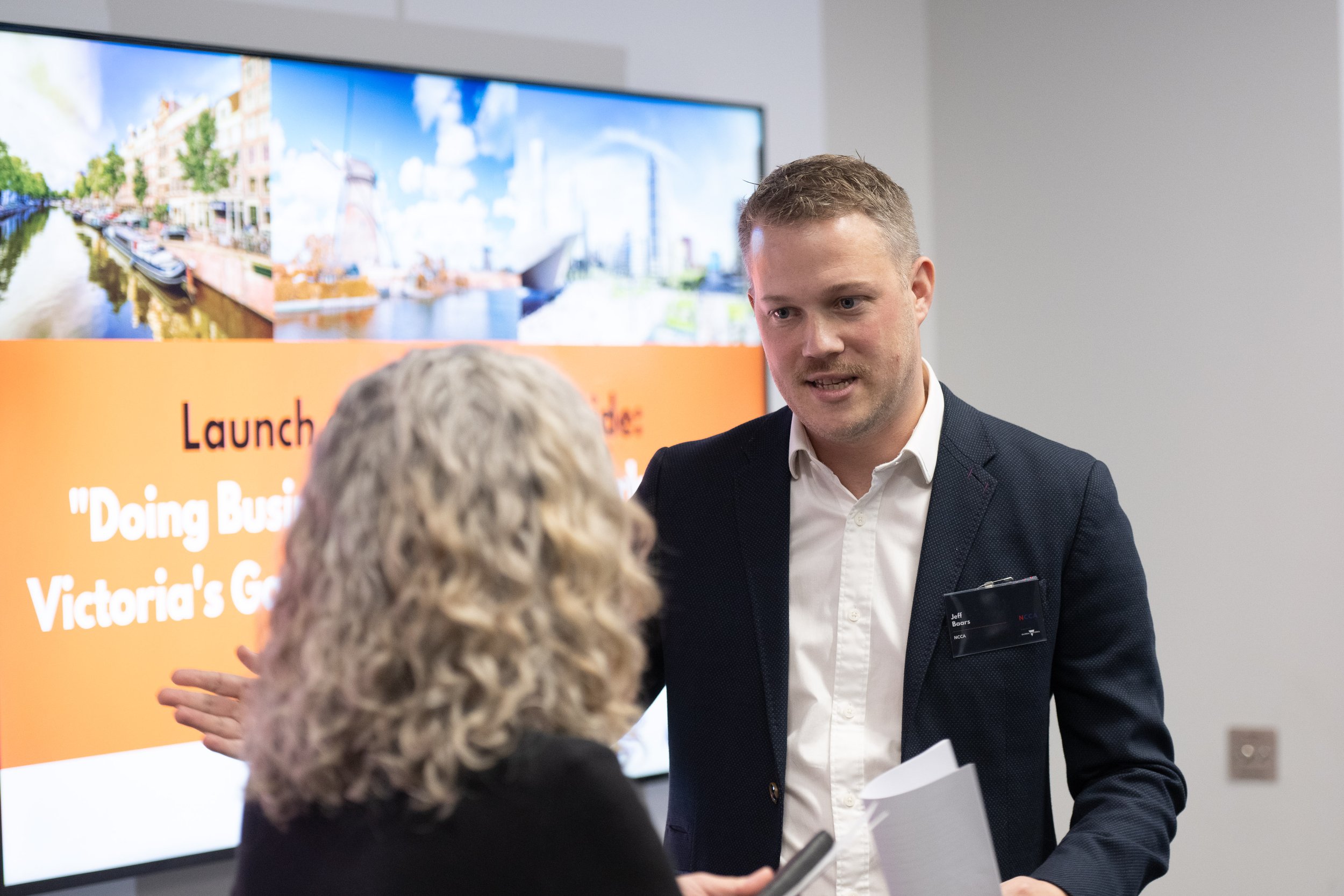 Man in navy blazer speaking to woman with gray curly hair in front of a presentation screen.