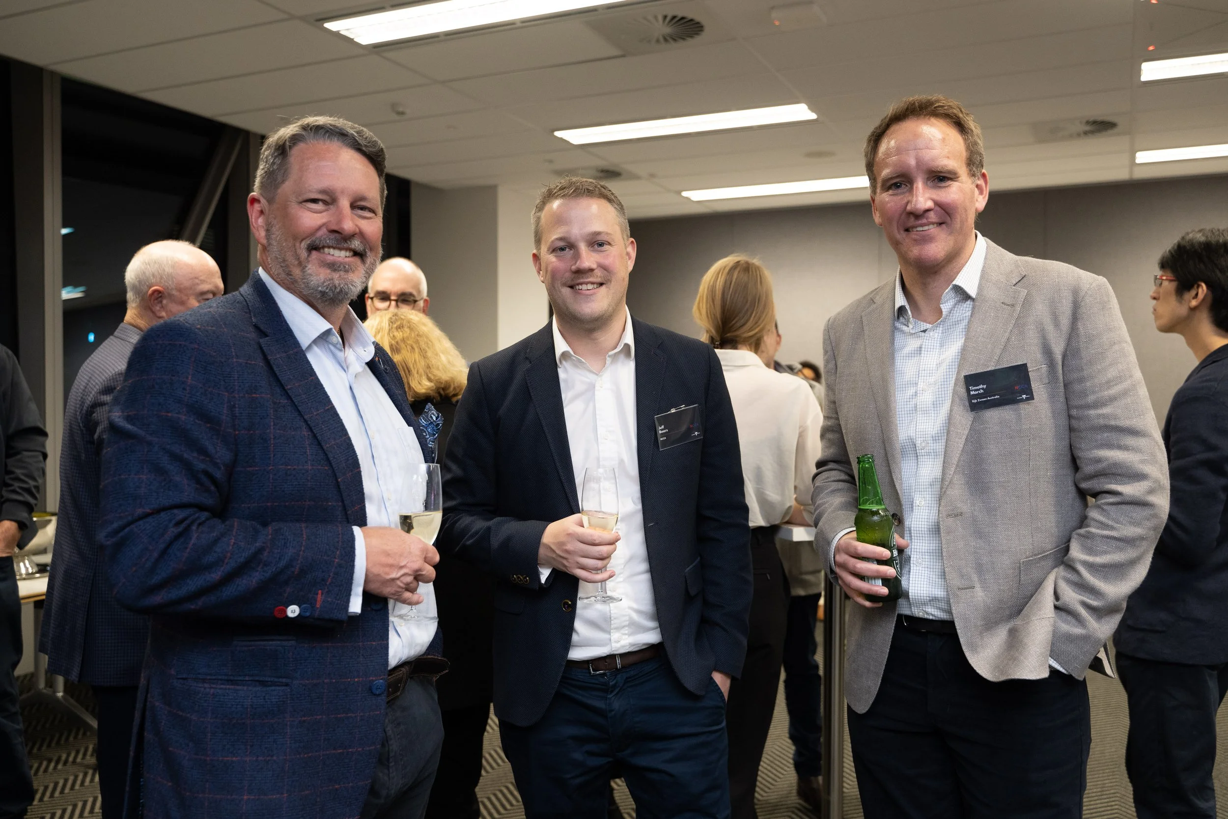 Three men in suits holding drinks at a professional network event, smiling at the camera, with other attendees in the background.