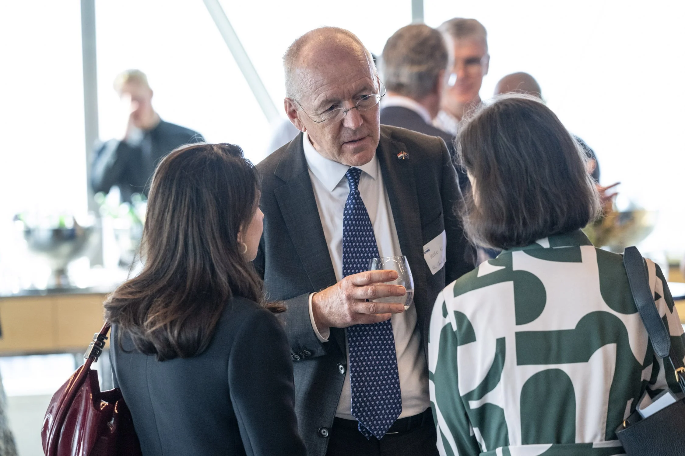 Three people engaged in conversation at a professional networking event, with a man holding a drink, wearing a suit and tie, and two women dressed in business attire.