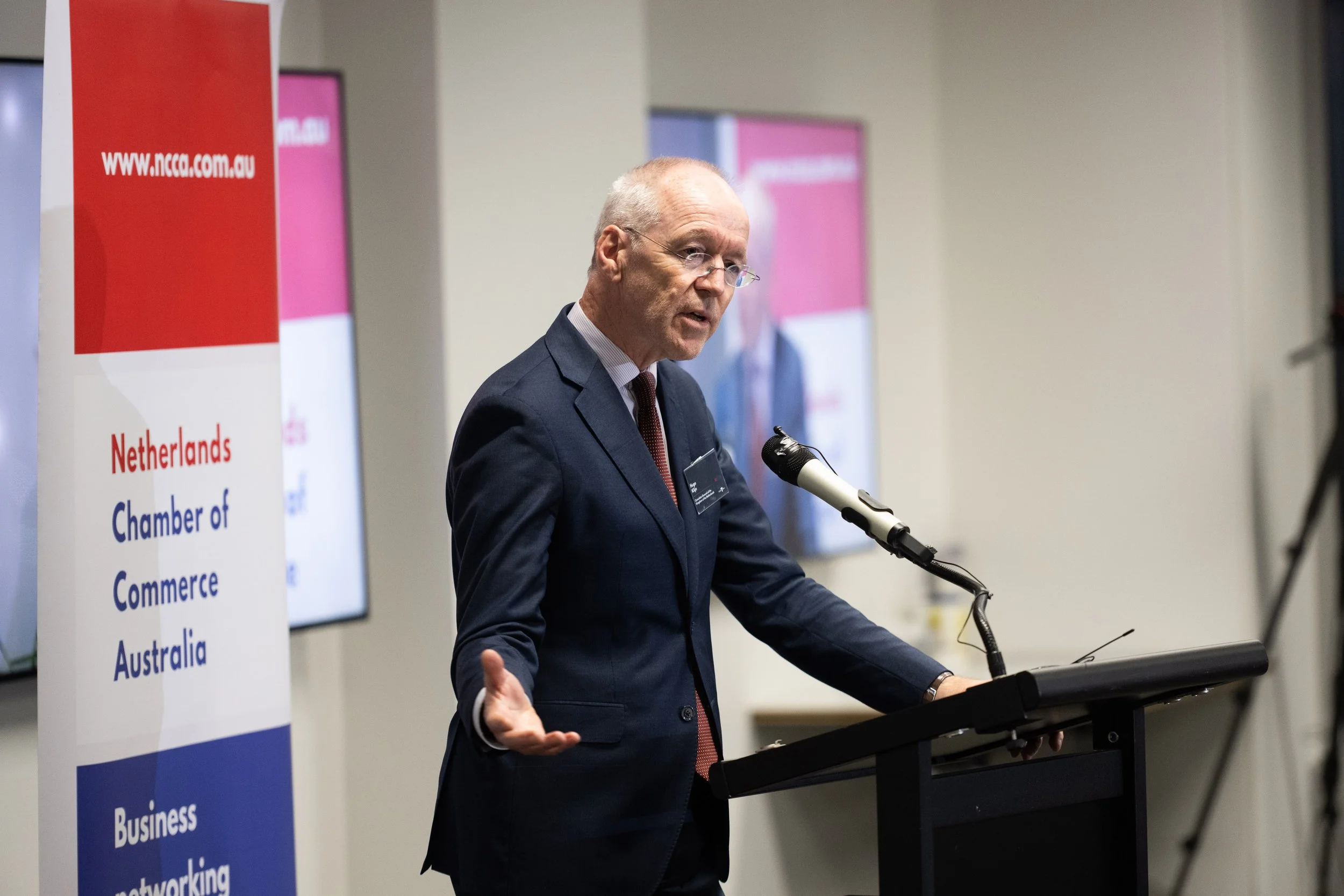A man in a suit speaking at a podium during a conference, with a banner for the Netherlands Chamber of Commerce Australia to his left.