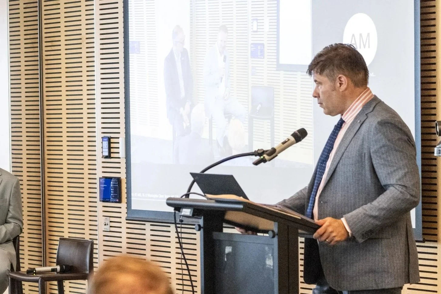 A man in a gray suit and striped shirt giving a presentation at a podium with a laptop and microphone in a conference room. There is a large screen behind him showing a projection of two other men.