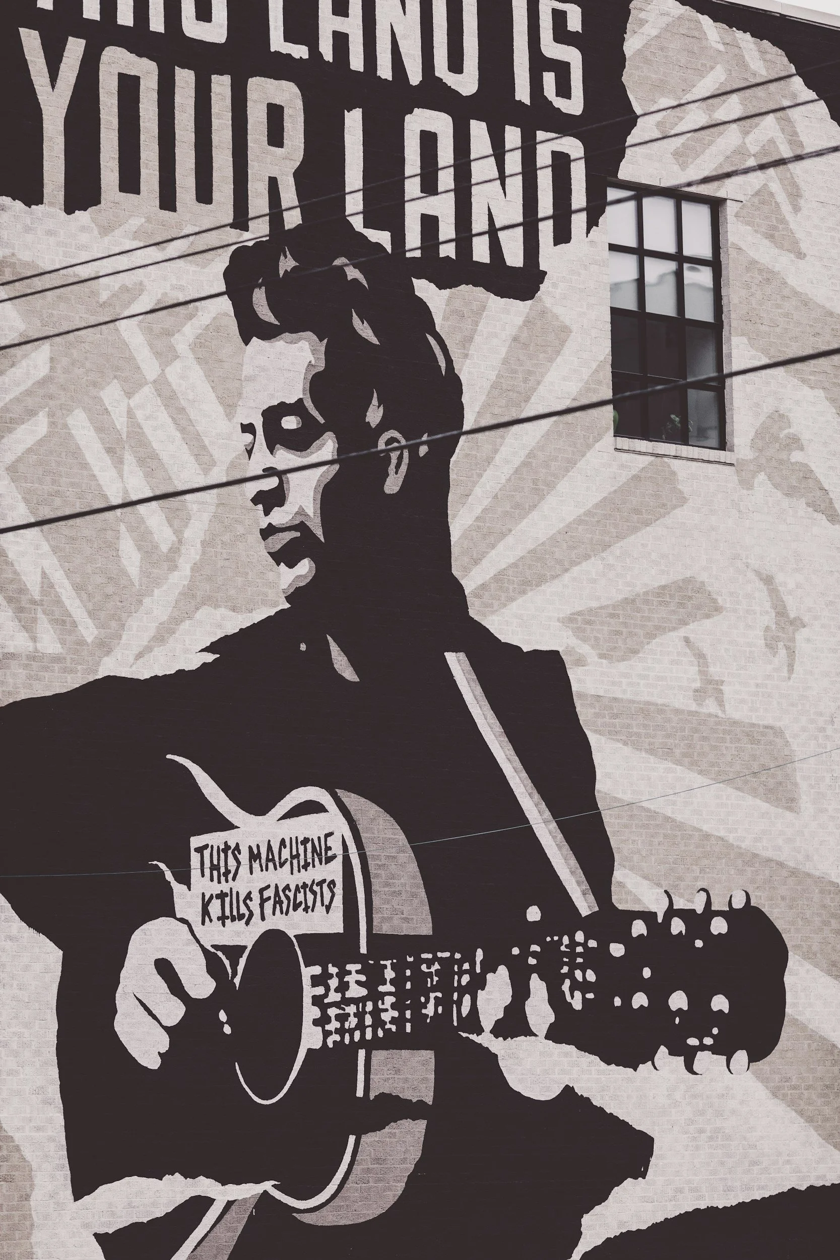 Street art mural of a folk artist with his hair in a pompadour playing an acoustic guitar. Text on the mural reads 'PHOTO LAND IS YOUR LAND' at the top and 'THIS MACHINE KILLS FASCISTS' on the guitar.
