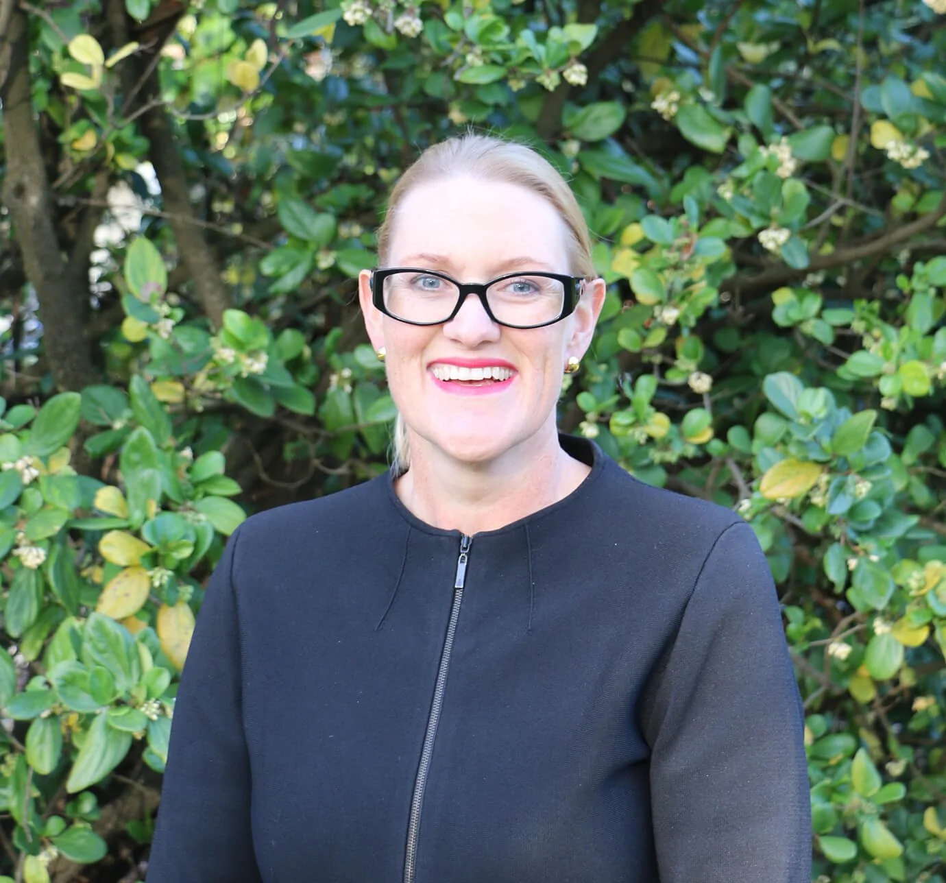 A woman with blonde hair, wearing black glasses and a black zip-up jacket, smiling in front of a bush with green leaves and white flowers.