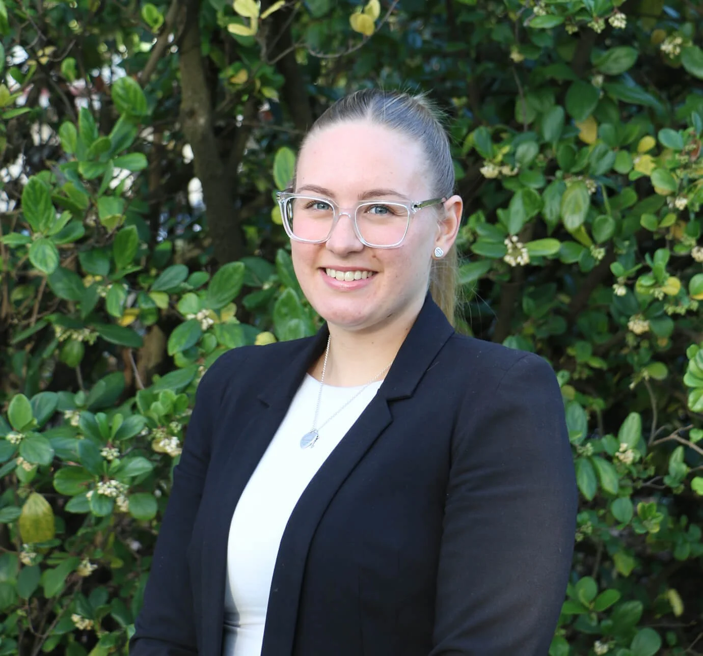 Portrait of a young woman with blonde hair tied back, wearing glasses, a black blazer, and a white top, standing outdoors in front of green foliage.