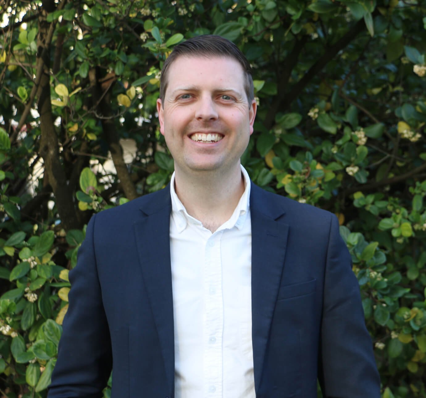 A smiling man in a white shirt and navy blazer standing outdoors in front of green foliage.