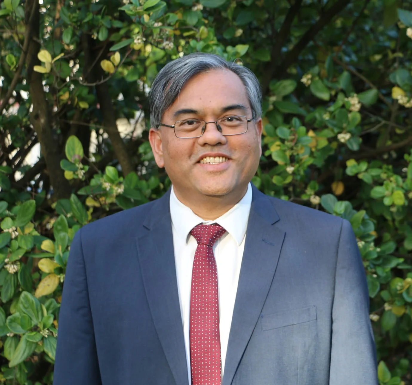 A man in a navy suit, white shirt, and red tie standing outdoors in front of green foliage, smiling at the camera.