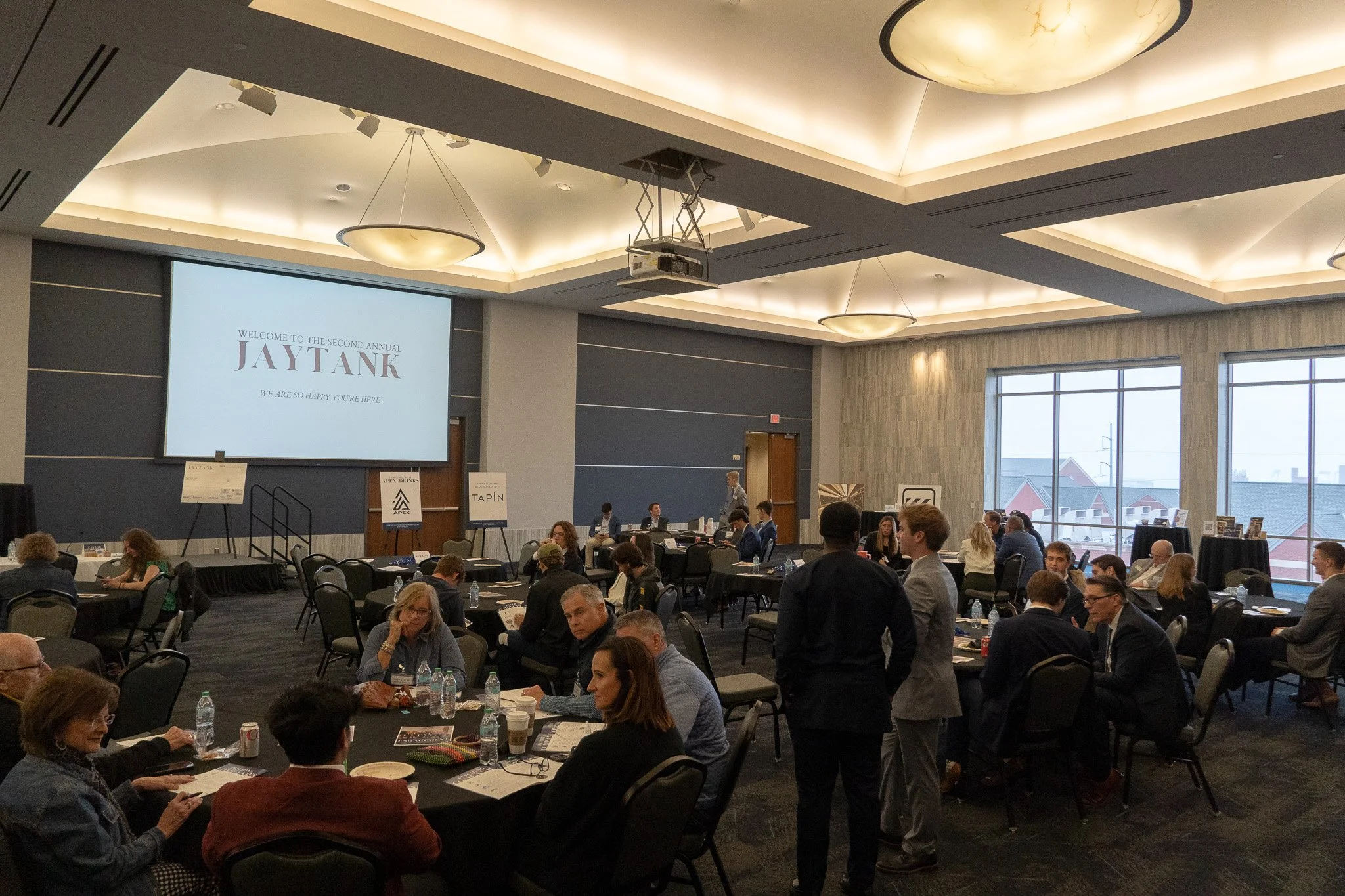A large conference room filled with round tables and people in business attire attending a meeting or event. There is a large screen at the front displaying a welcome message for the second annual Jay Tank event. Windows on the right side show an outdoor view of rooftops and cloudy weather.