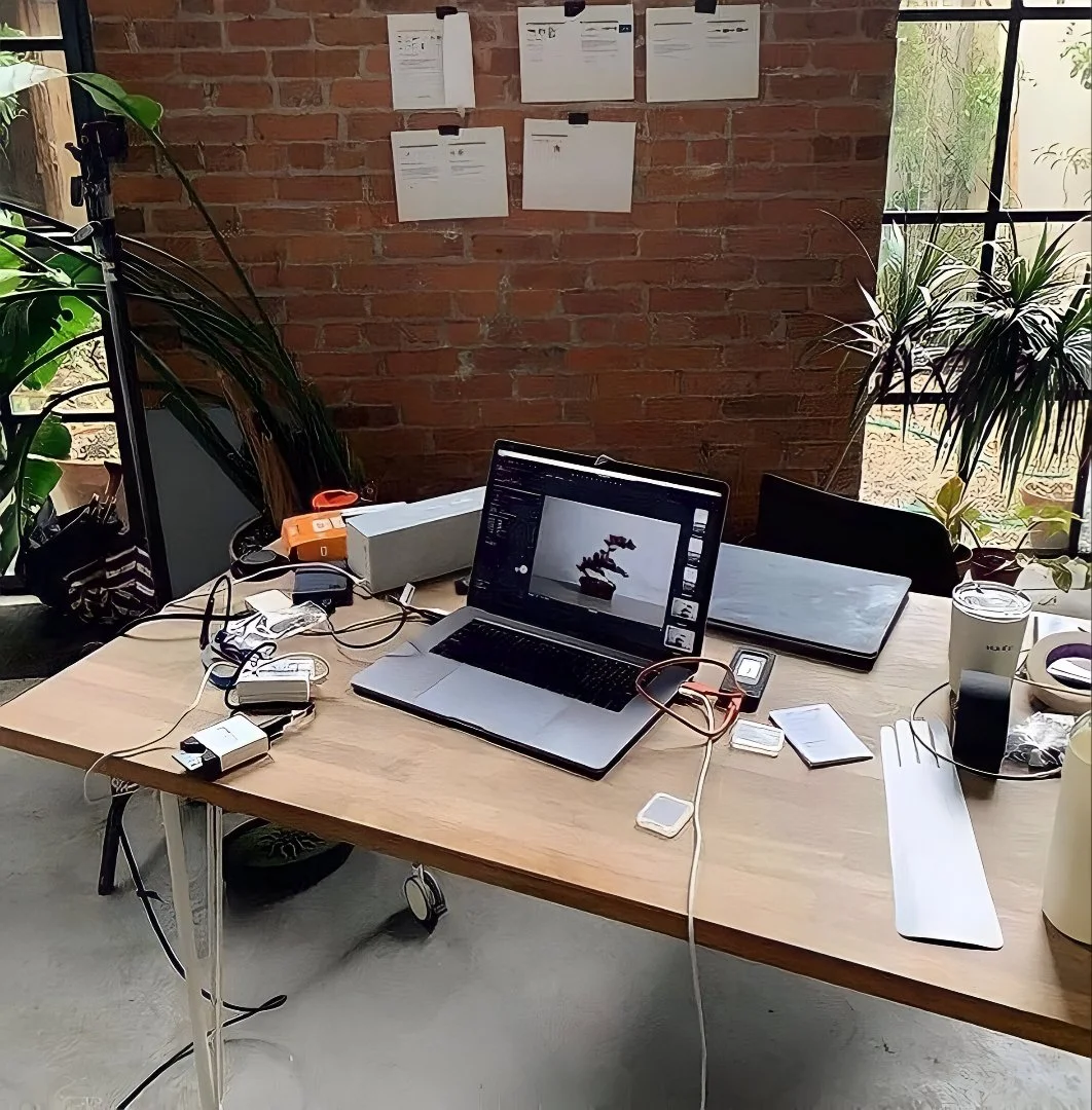 A cluttered workspace with a wooden desk, two laptops, various electronic accessories, and potted plants near large windows.