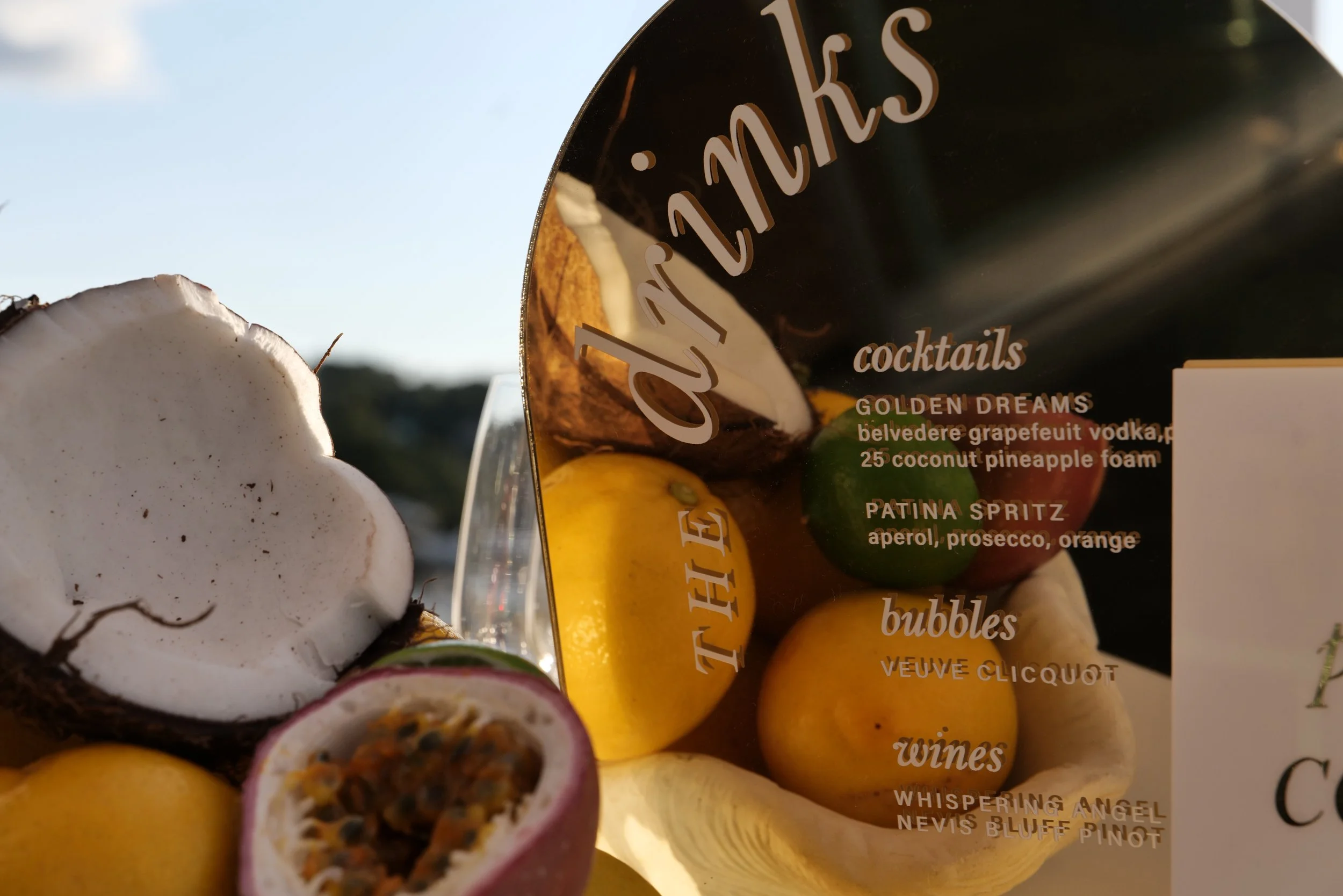 Close-up of a halved coconut and a box of cocktail ingredients featuring passion fruit, lemon, lime, and other fruits against a blue sky background.