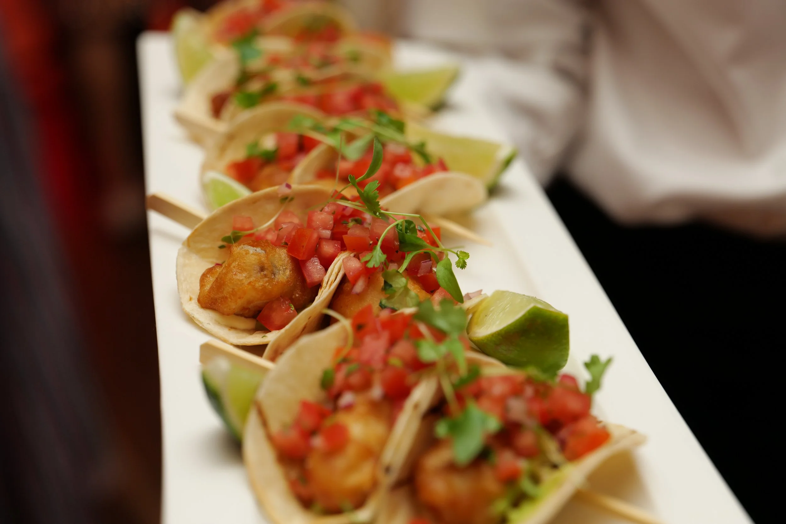 Close-up of a row of Mexican tacos topped with diced tomatoes, cilantro, and lime wedges on a white tray.