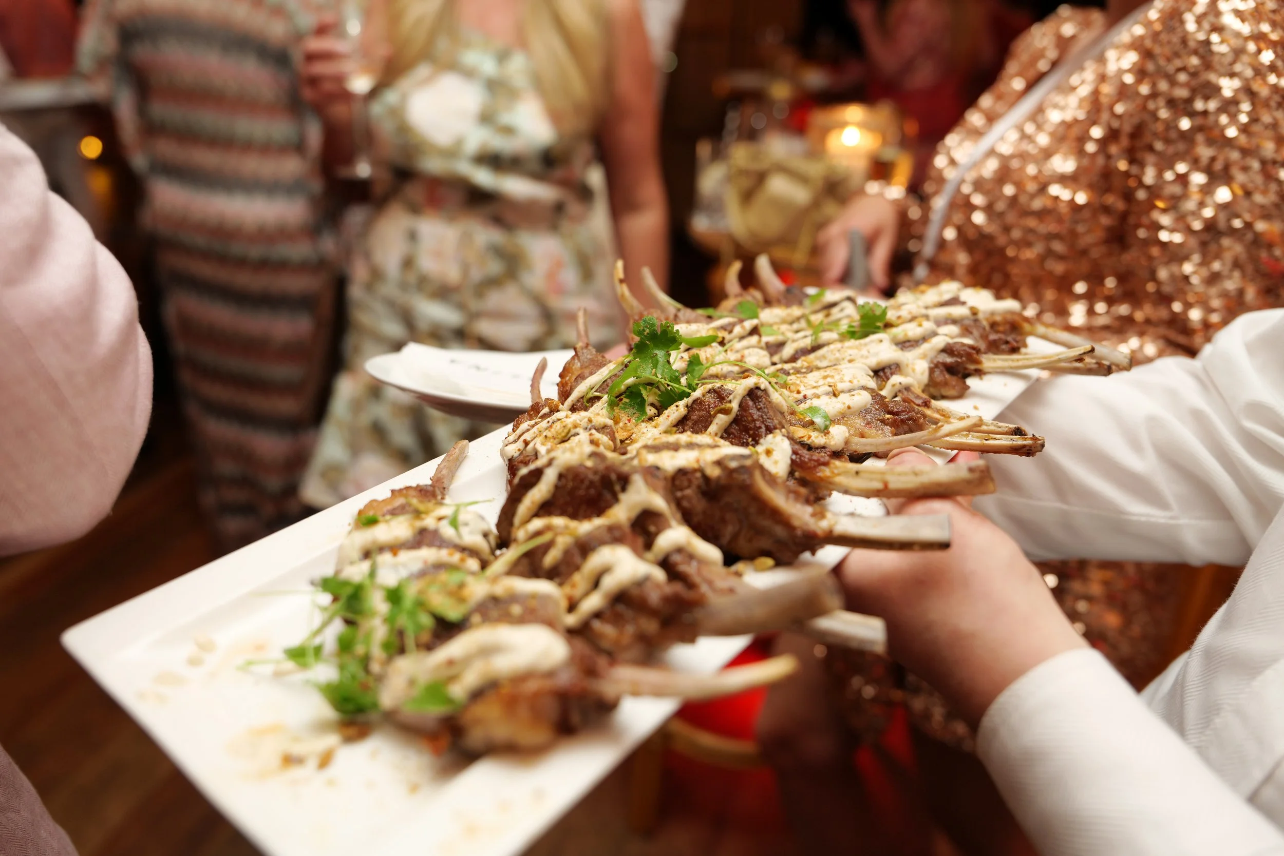 Hands holding a platter of grilled meat bones garnished with cilantro and drizzled with sauce at a festive gathering.