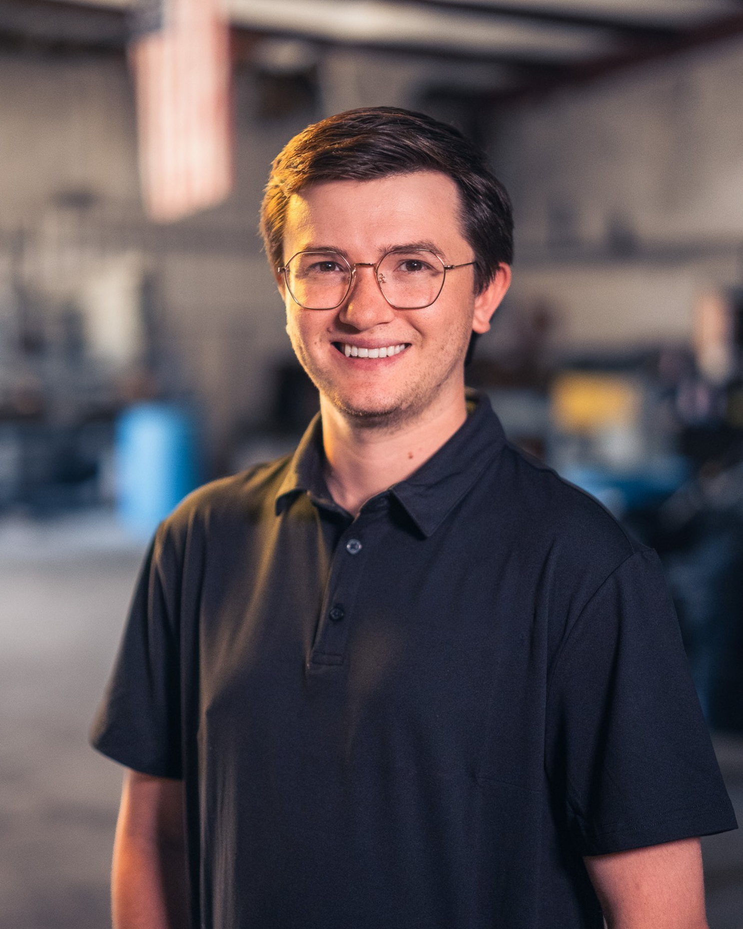 A young man with glasses and dark hair smiling at the camera in a workshop or garage setting.