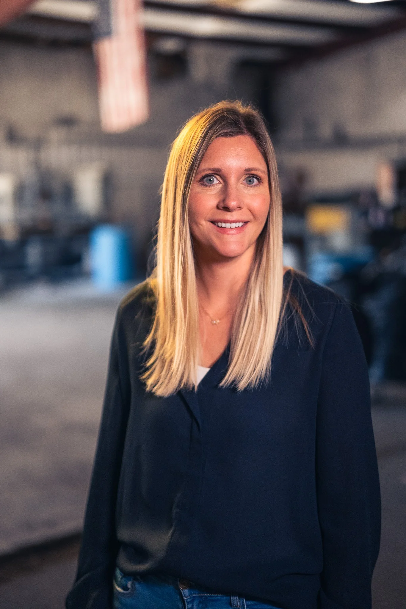 A woman with long blonde hair smiling indoor, blurred background.