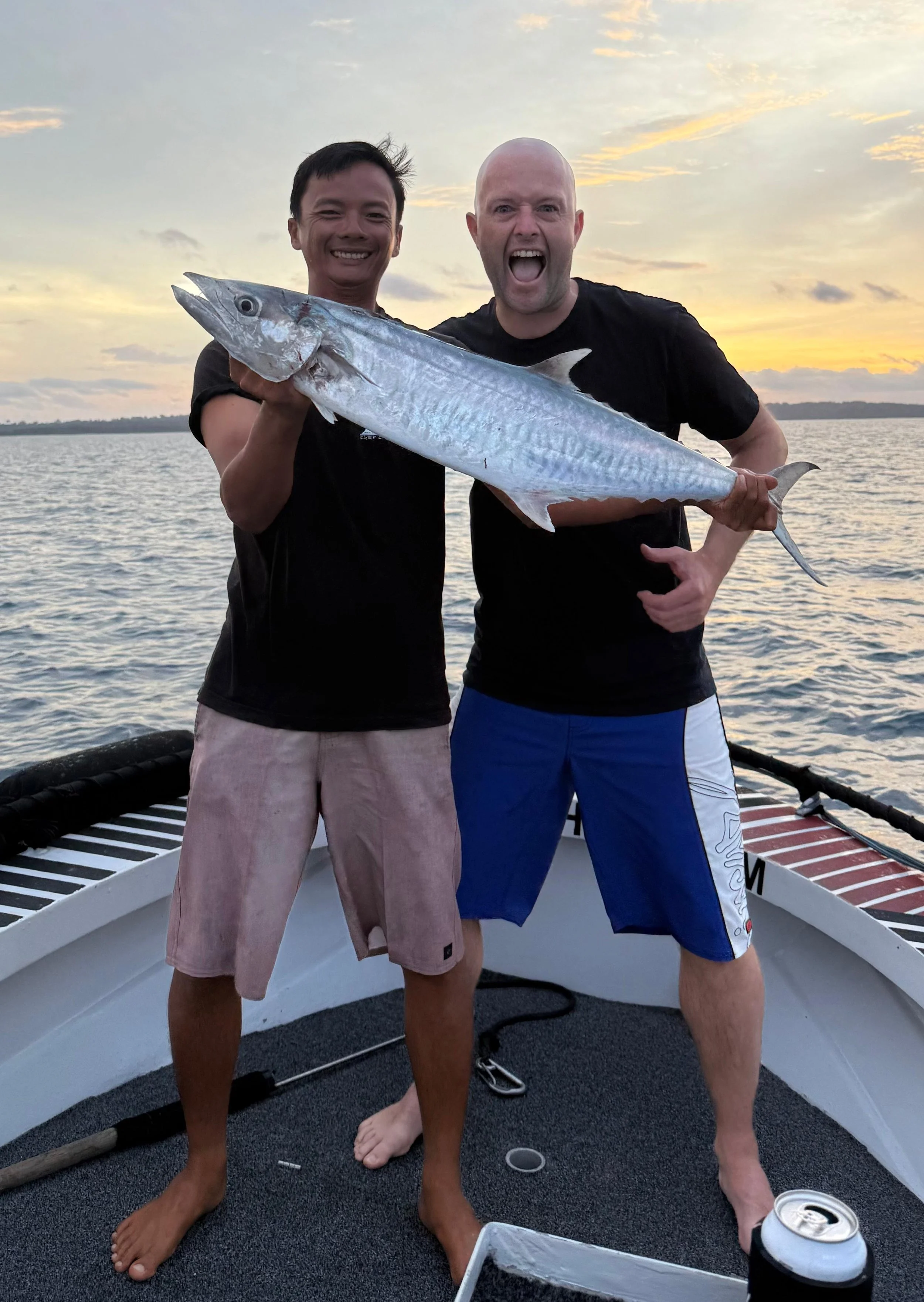 Two men on a boat holding a large fish, with a sunset over the water in the background.