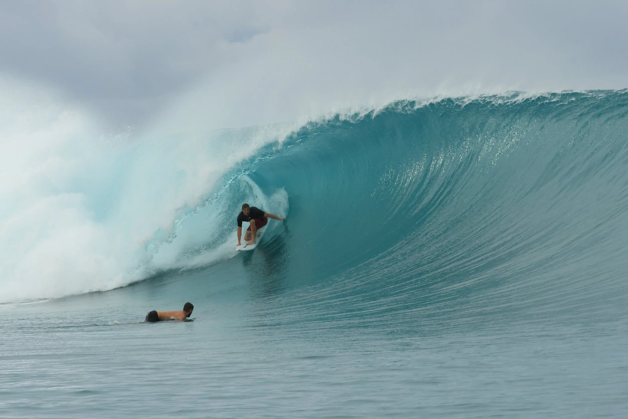 A person surfing on a large blue wave while another person paddles in the water nearby.