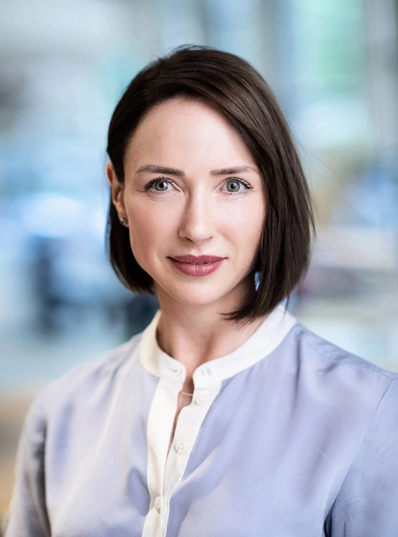 Portrait of a woman with shoulder-length dark brown hair and blue eyes, wearing a light-colored blouse, in a blurred indoor setting.