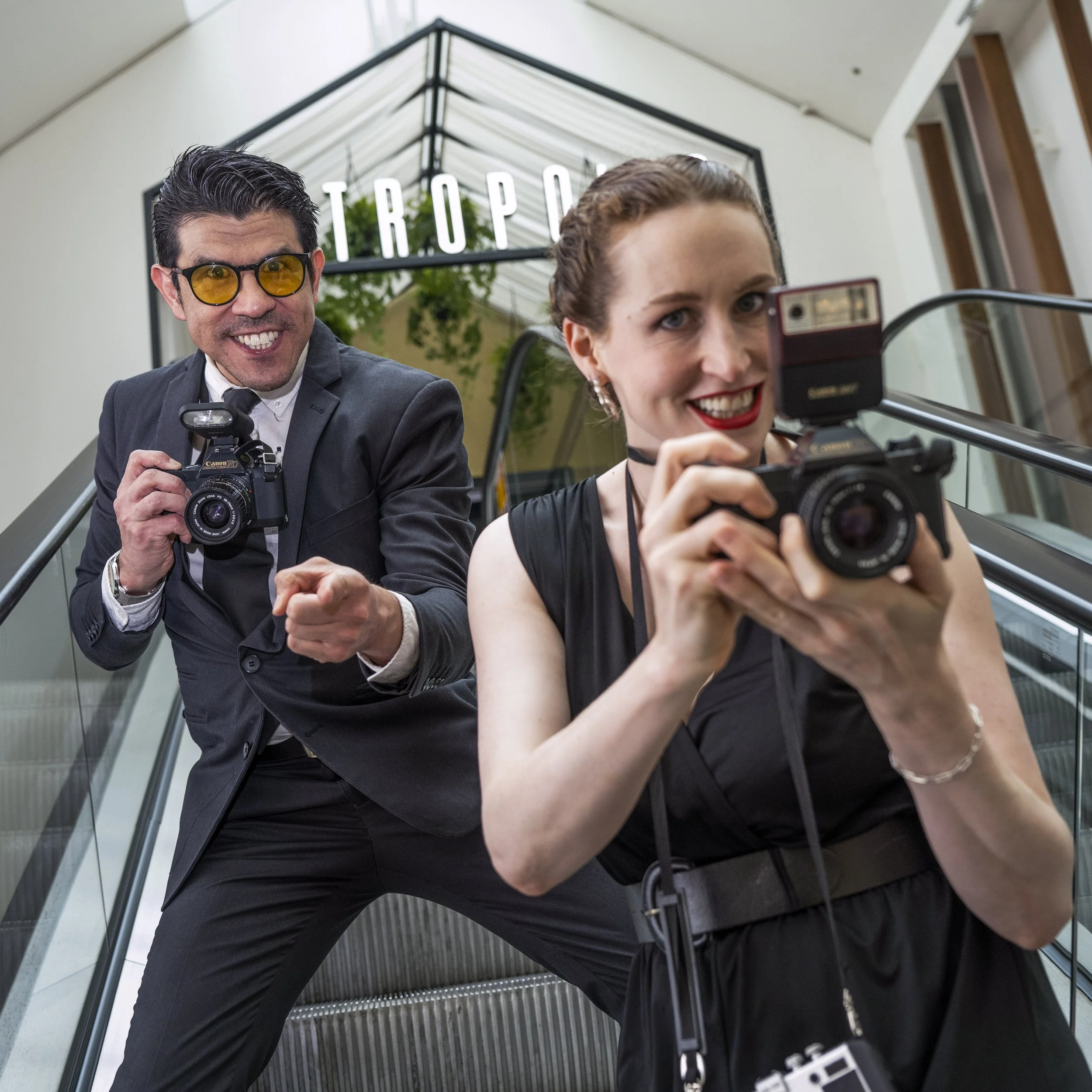 Two people, a woman and a man, are on an escalator taking a selfie. The woman holds the camera and smiles at it, wearing a black dress, and the man stands behind her in a suit and tie, pointing and smiling. There is a sign that says 'TROPO' in the ba