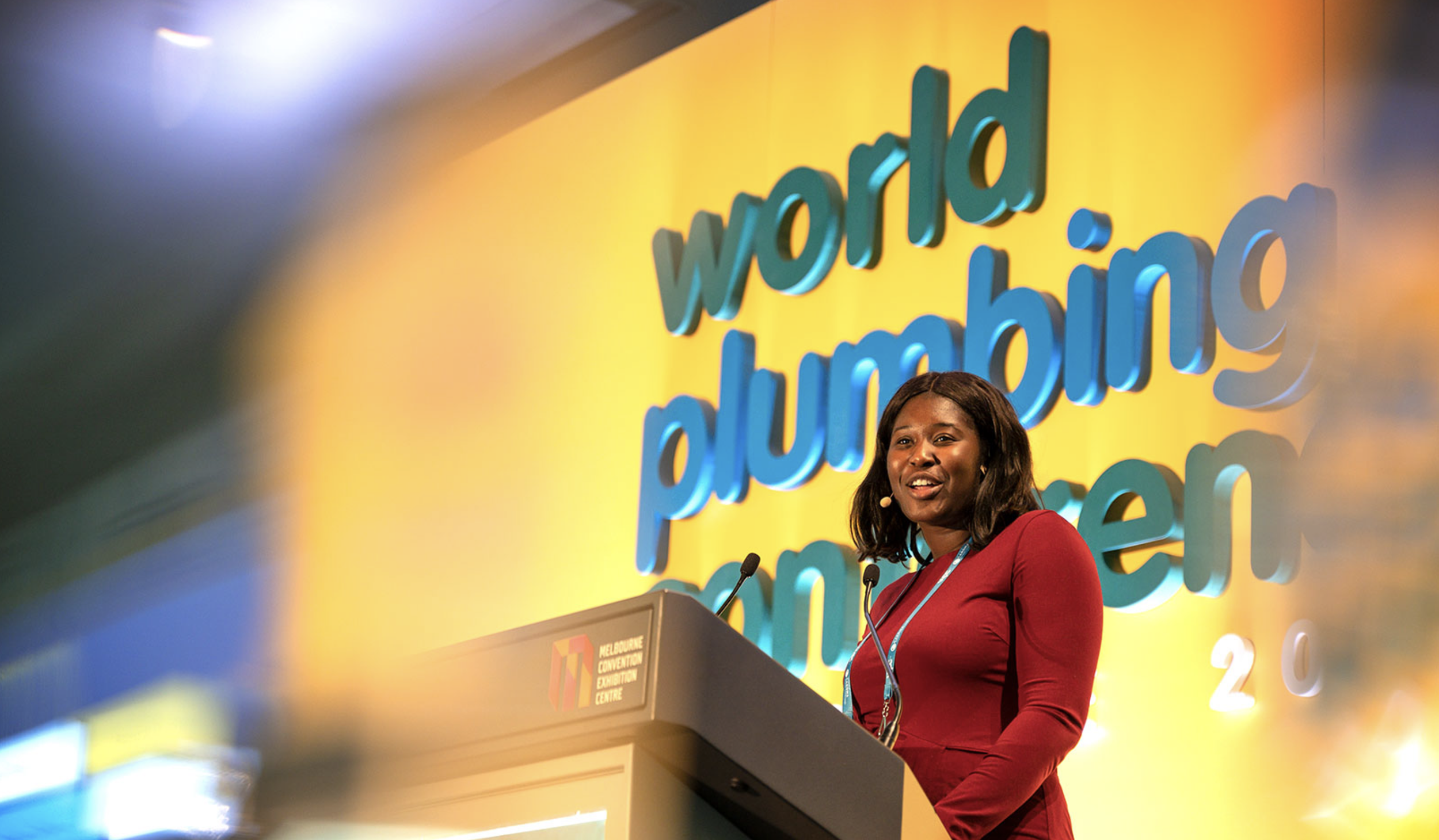 A woman speaking at a podium during the Melbourne Convention and Exhibition Centre conference, with a large yellow backdrop featuring the words 'world unfolding'. she is wearing a red dress and has a microphone headset.