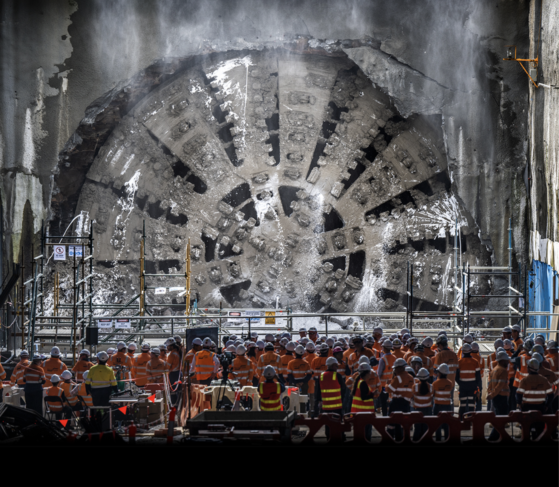 Construction workers in orange safety attire and white helmets gathered in a tunnel with a large circular tunnel boring machine behind scaffolding.