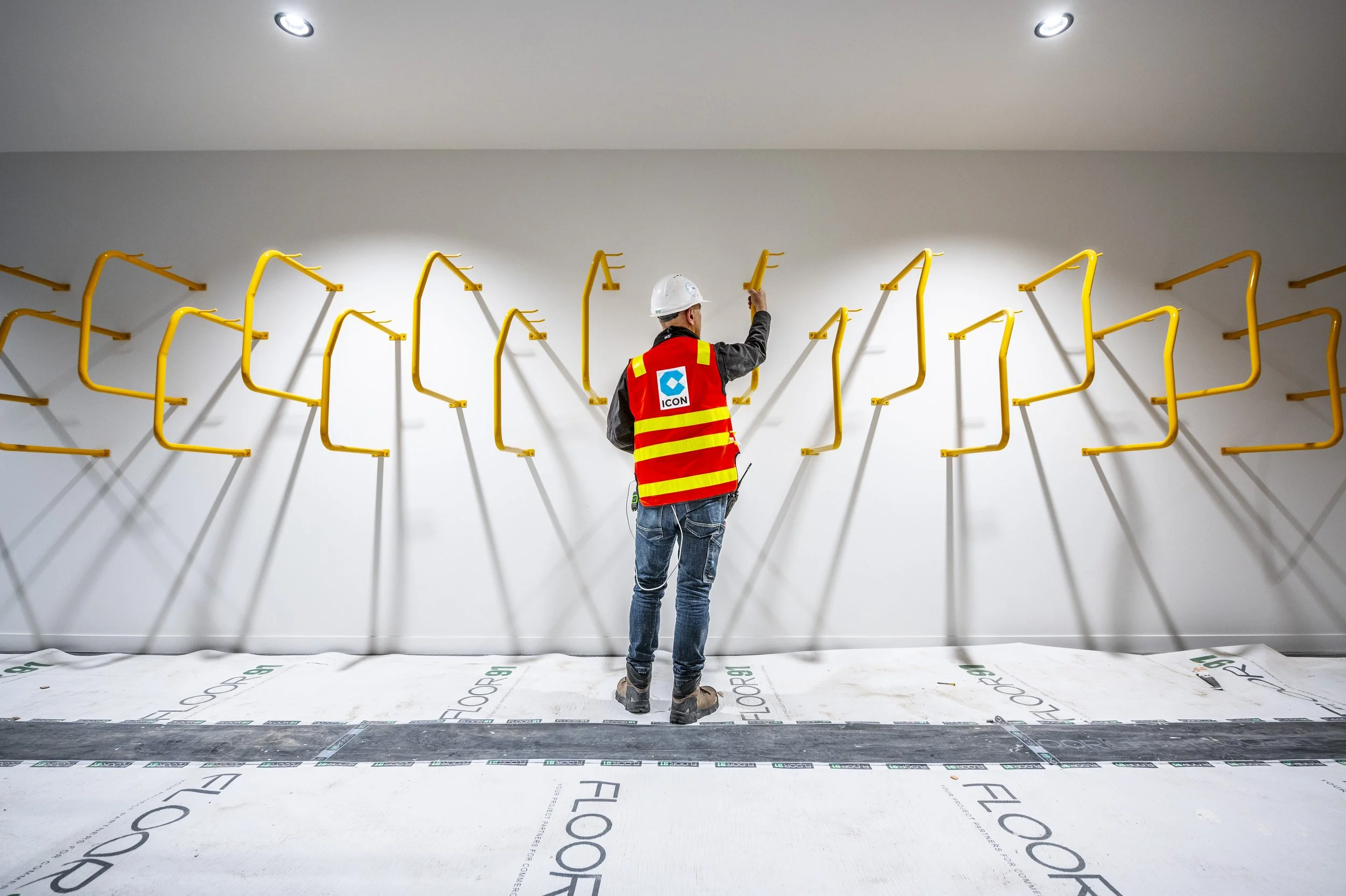 A construction worker wearing a white safety helmet and a red high-visibility vest with reflective yellow stripes operates on an indoor wall with yellow pipes arranged in a pattern. The floor is covered with protective sheeting marked 'FLOORProtectio