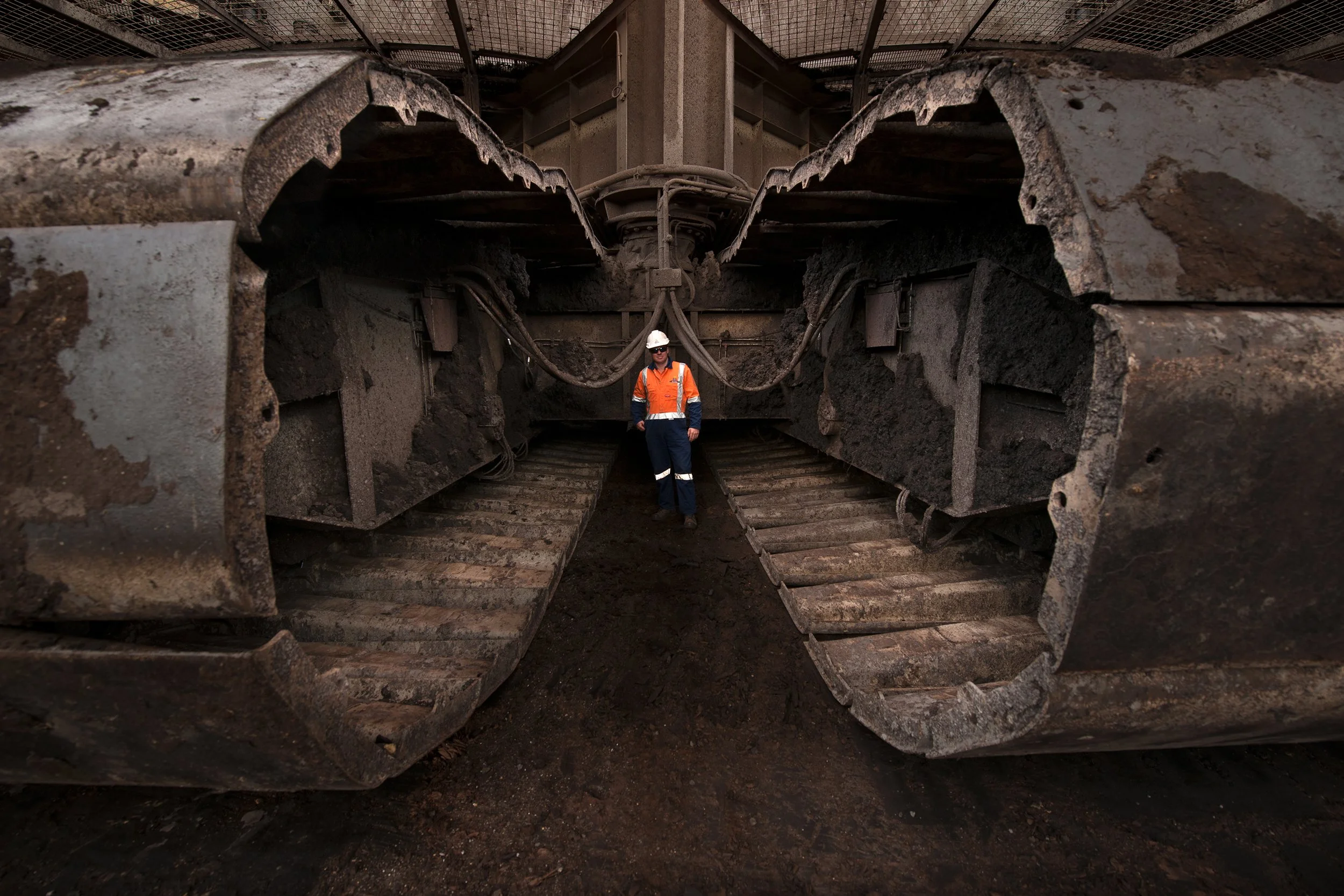 A worker in safety gear, including a white hard hat, orange reflective vest, and dark blue overalls, standing between large, dirty tracks of a construction or mining vehicle underground.