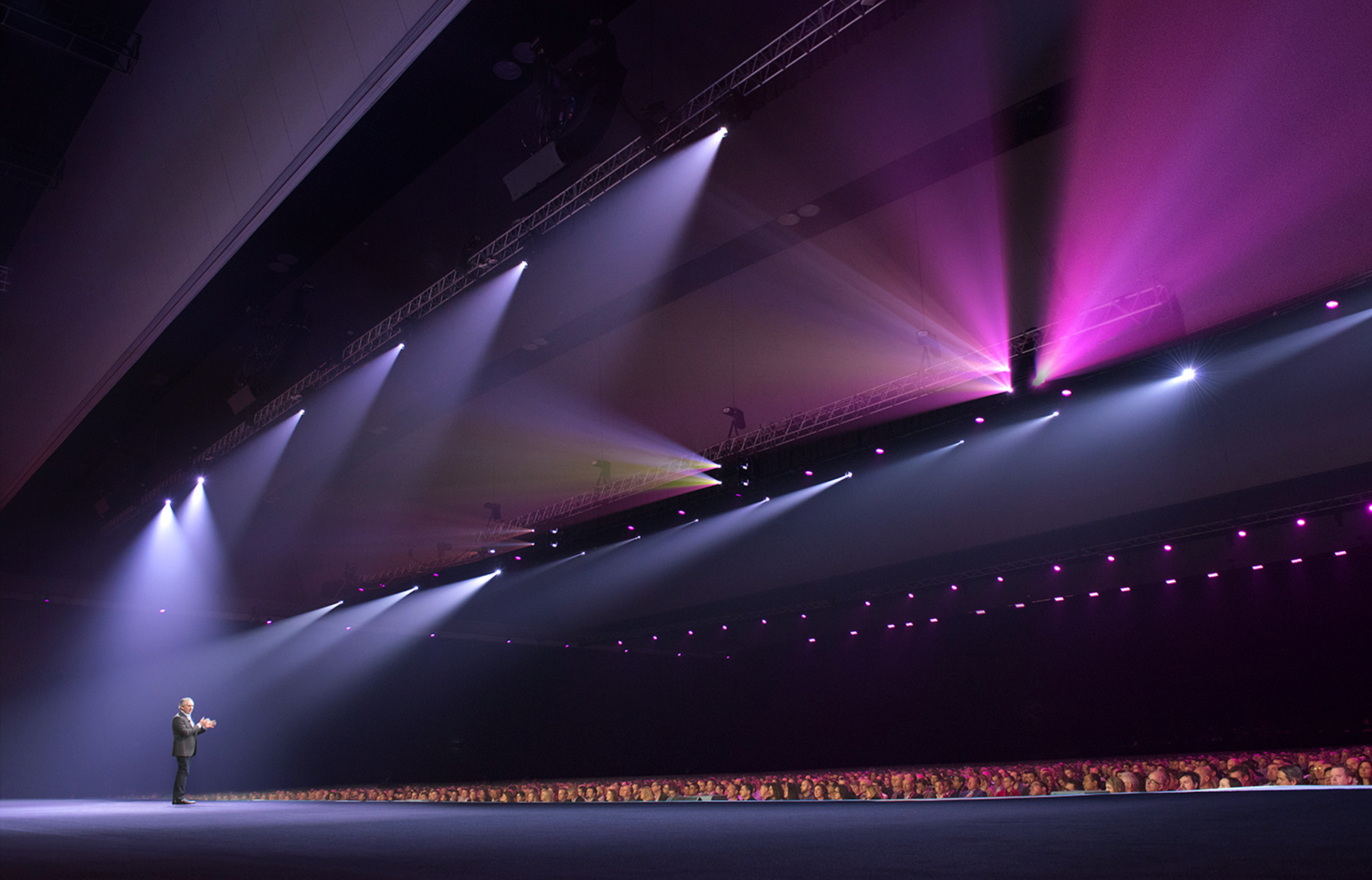 A speaker on stage in a large auditorium with bright spotlights, purple and white stage lighting, and an audience.