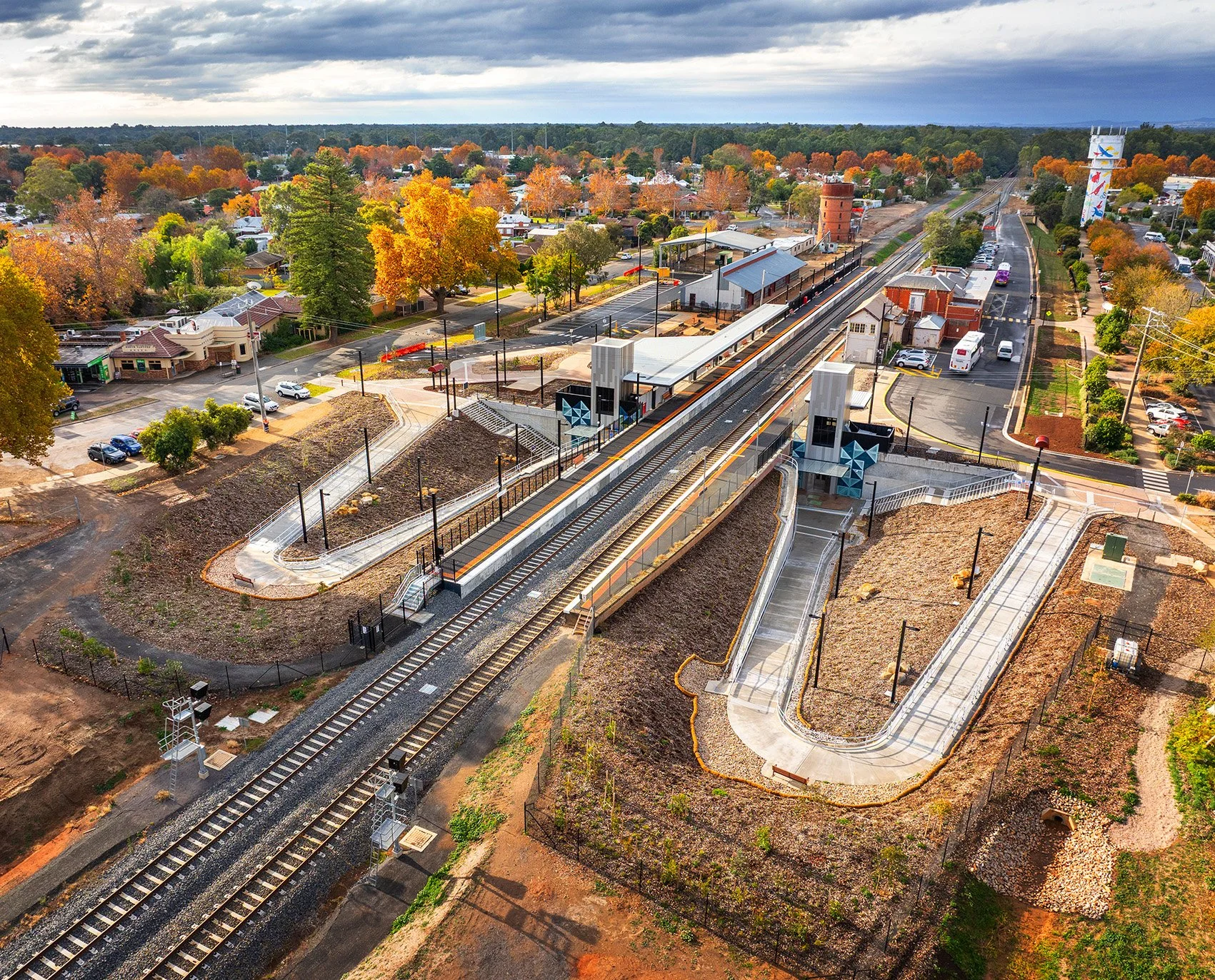 Aerial view of a modern train station under construction with multiple railway tracks, surrounded by trees with autumn foliage, parking lots, and a small town in the background.