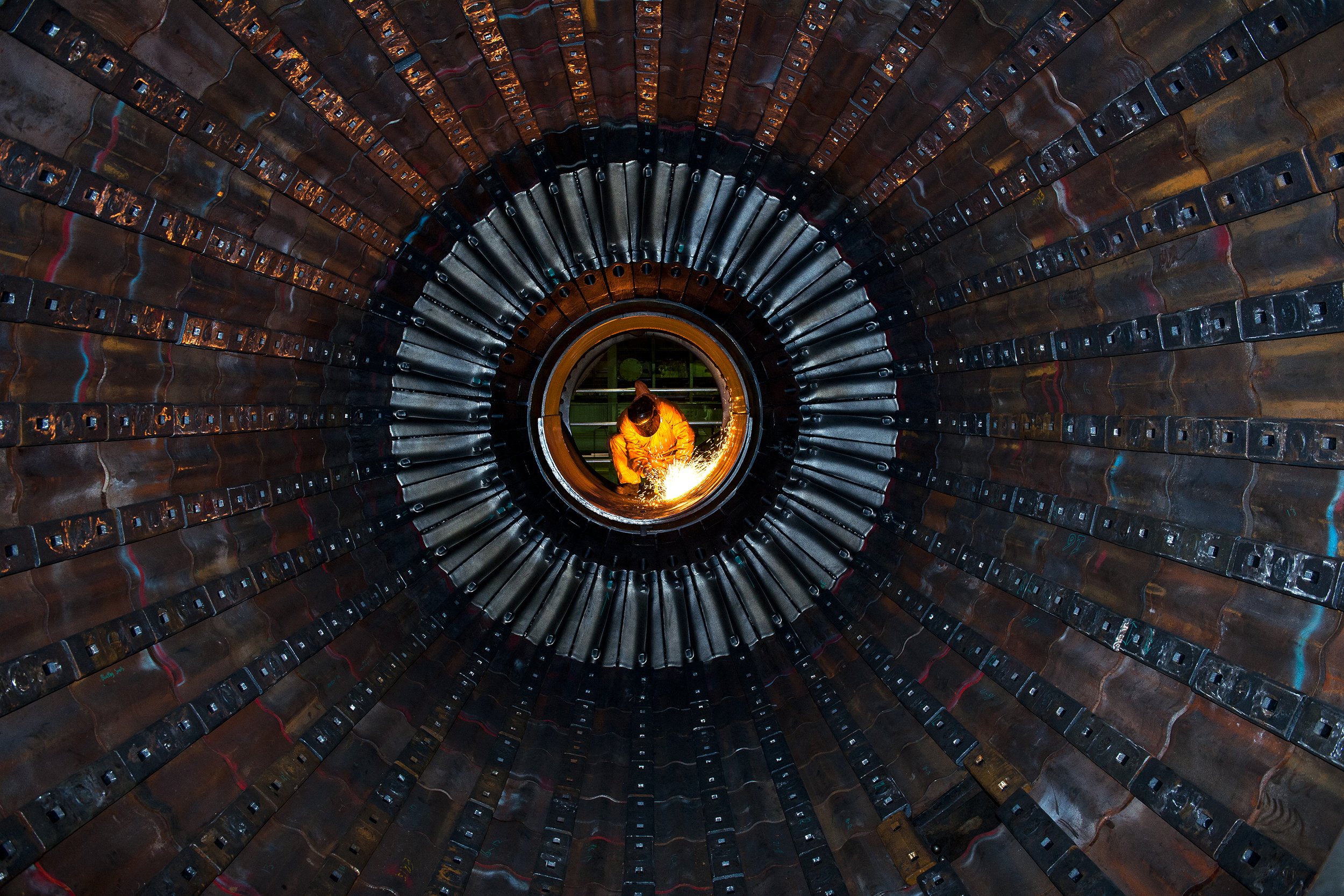 A worker in a yellow protective suit welding inside a large metal pipe or tunnel, with sparks flying.