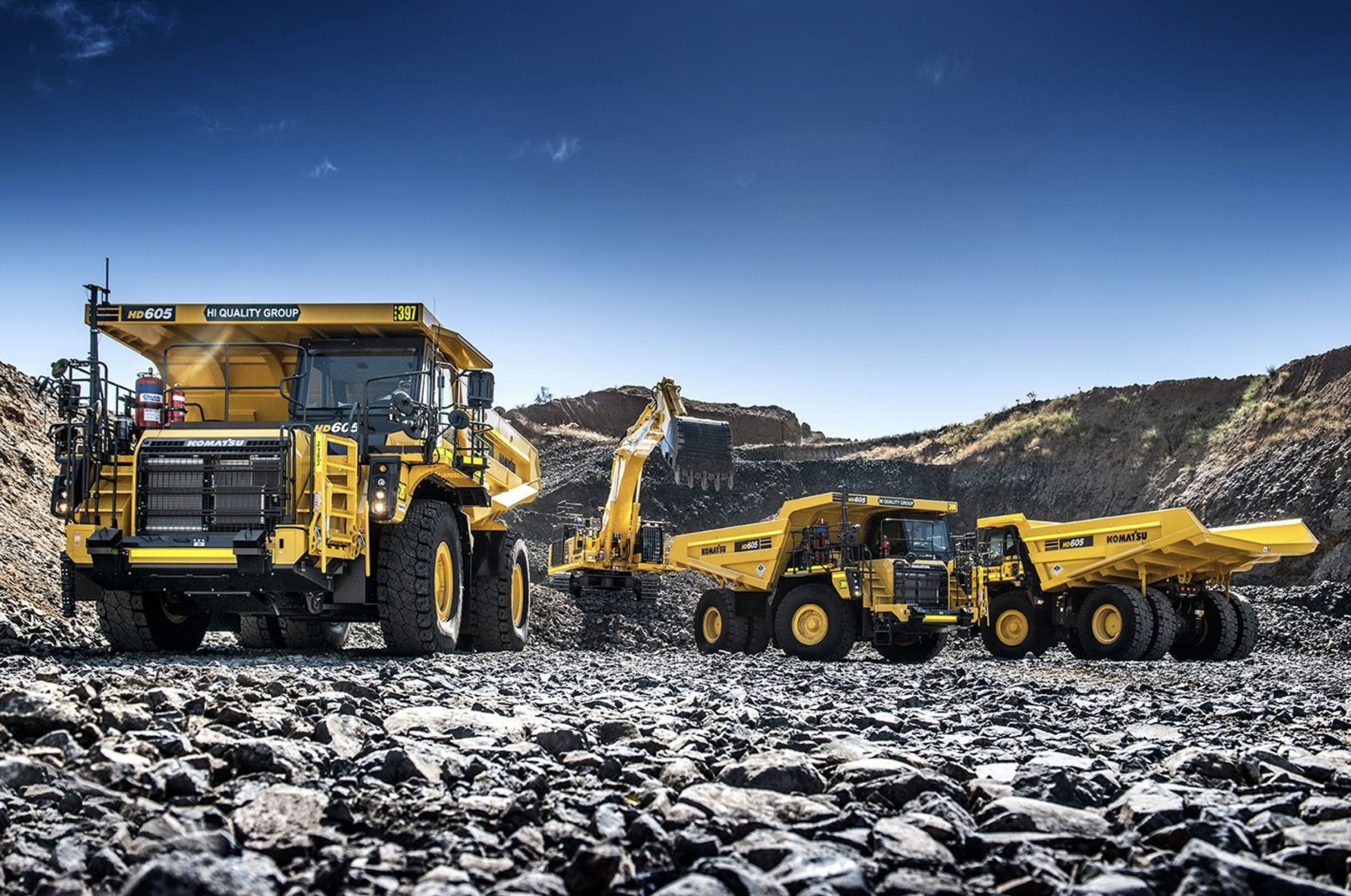 Three large yellow construction trucks working on a rocky construction site under a clear blue sky.