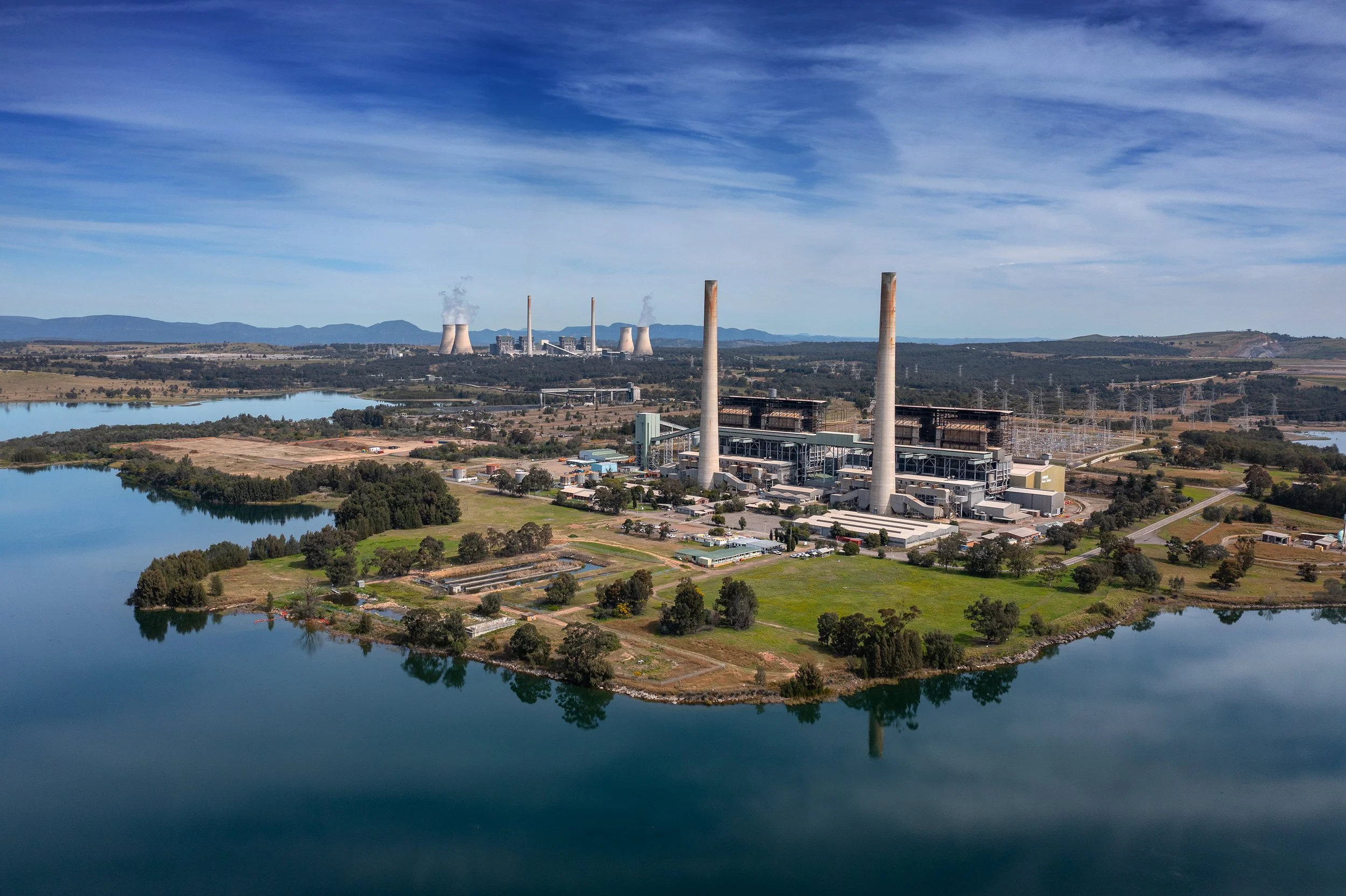 Aerial view of a large industrial power plant with cooling towers and smokestacks, surrounded by lakes and green landscapes under a blue sky.