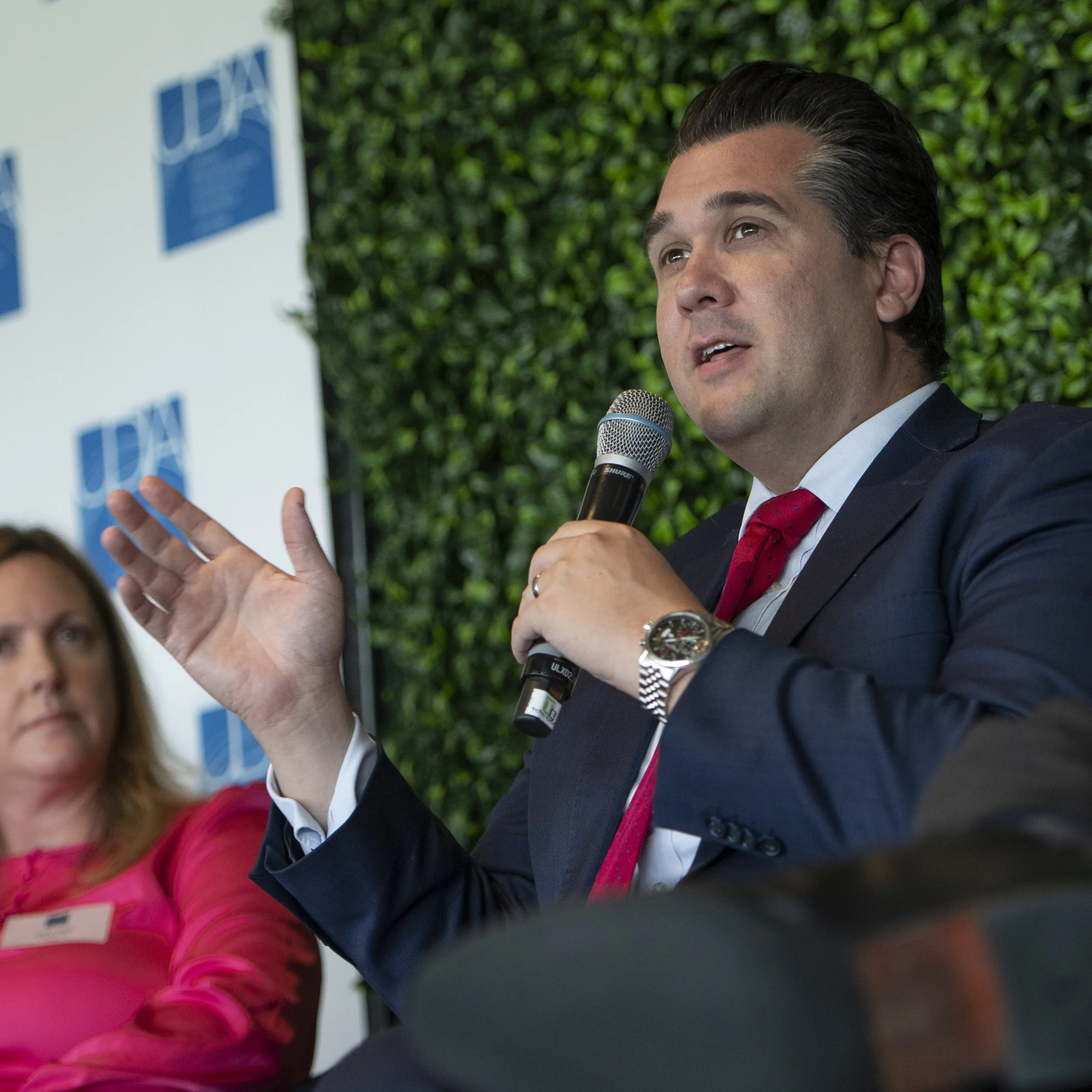 A man in a suit and red tie speaking into a microphone during a panel discussion, with a woman in a pink blouse sitting beside him, and a background with greenery and a banner with the letters 'UFA'.