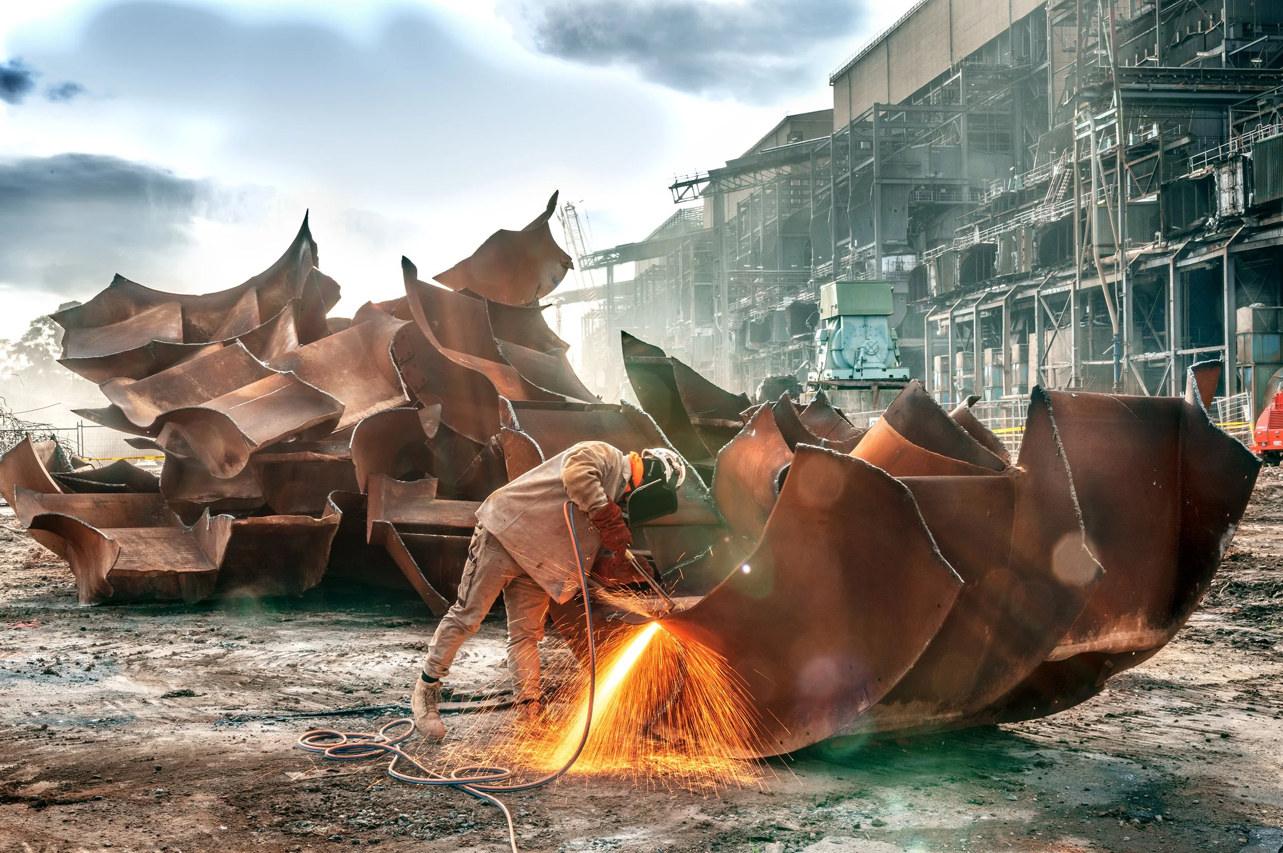 A worker welding a large, rusted metal structure outdoors with sparks flying, industrial buildings in the background, and a partly cloudy sky.