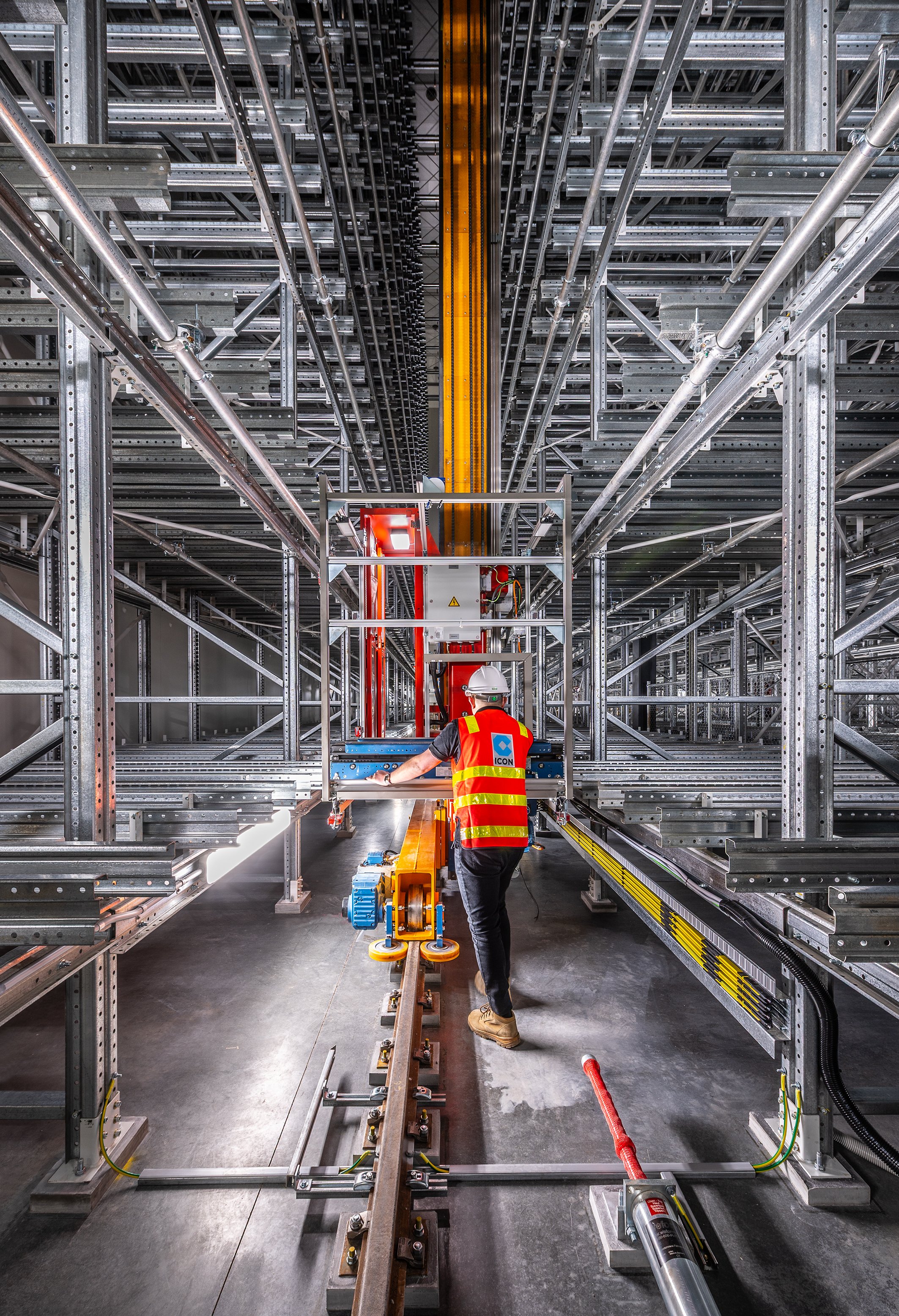 Construction worker operating machinery inside a large industrial warehouse with metal shelving and rails.