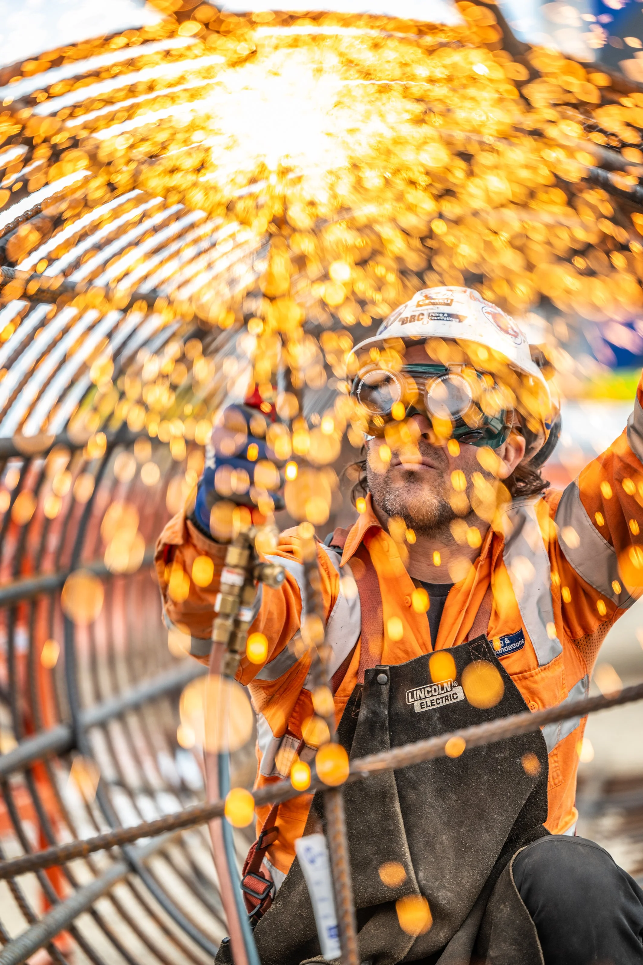 Worker welding metal rebar, sparks flying, safety gear including helmet and glasses, construction site background.