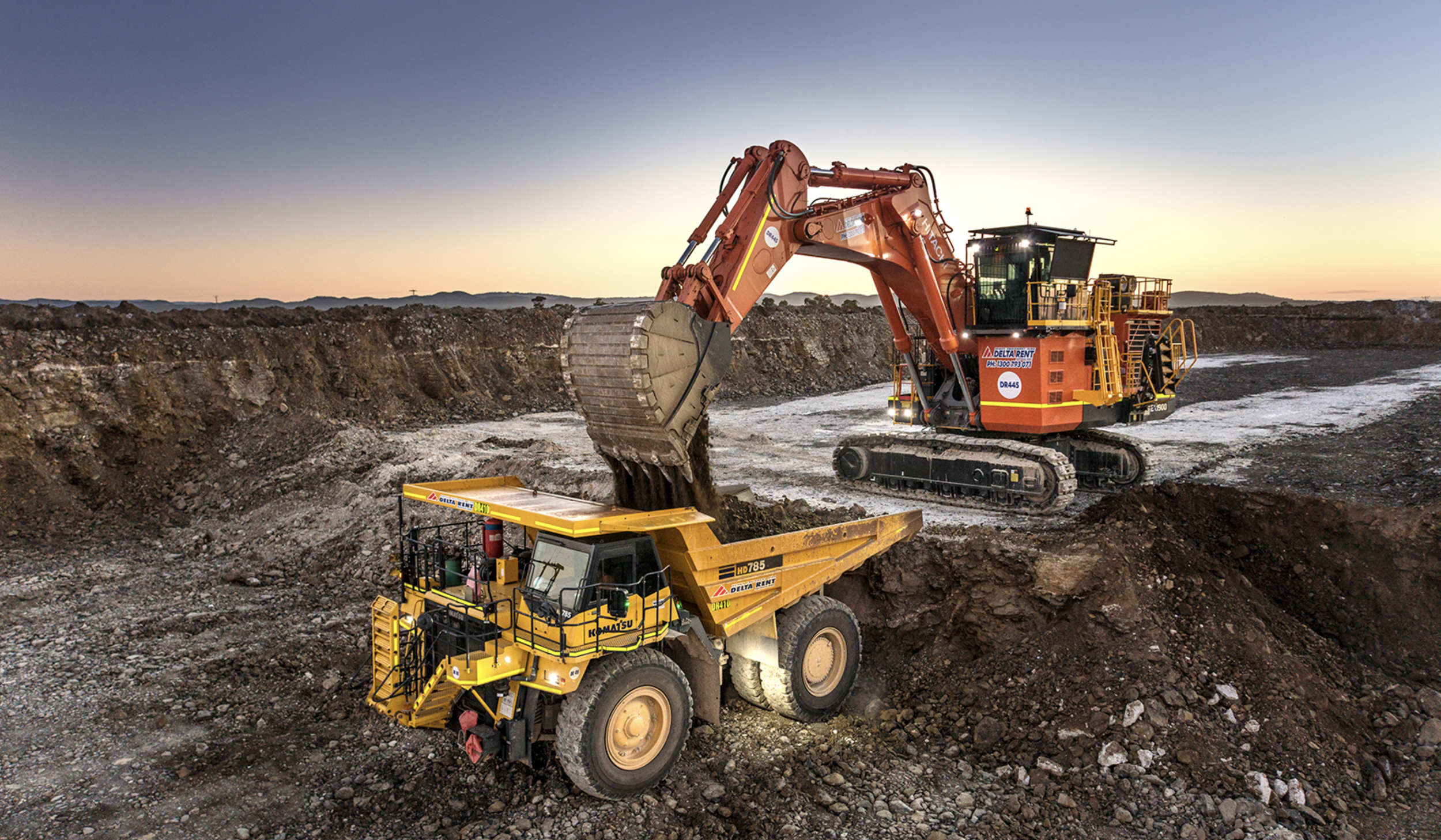 An excavator and a dump truck working together in a mining site at sunset.