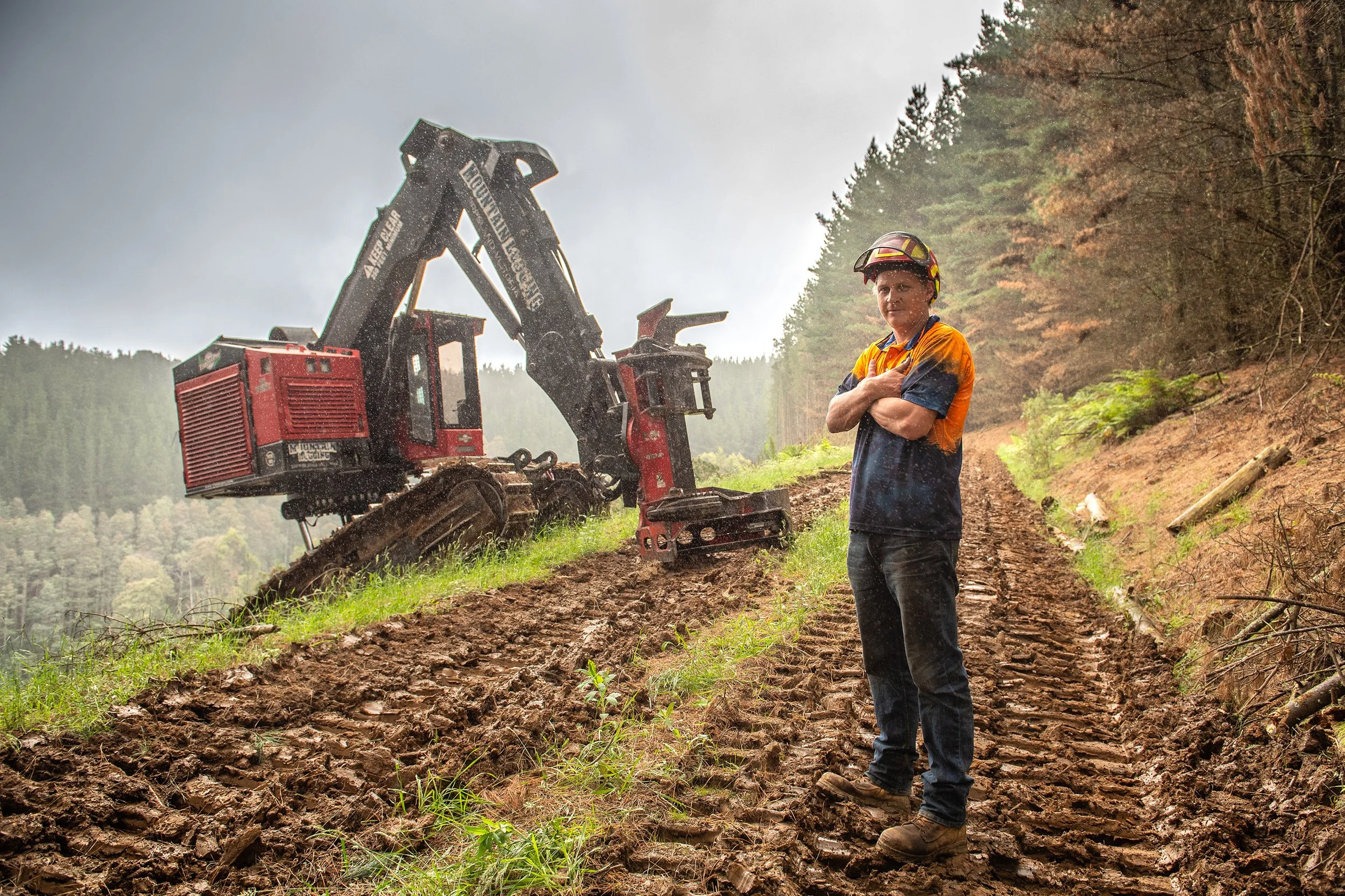 A person standing on a muddy trail wearing a helmet and orange and dark blue work clothes, with a large excavator in the background on a forested hillside during rainy weather.