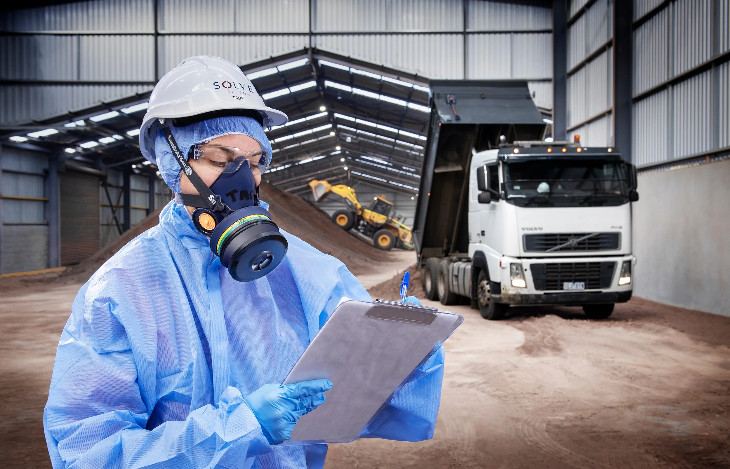 A worker in protective gear, including a mask, goggles, and a helmet, is writing on a clipboard inside an industrial warehouse with construction equipment and a dump truck in the background.