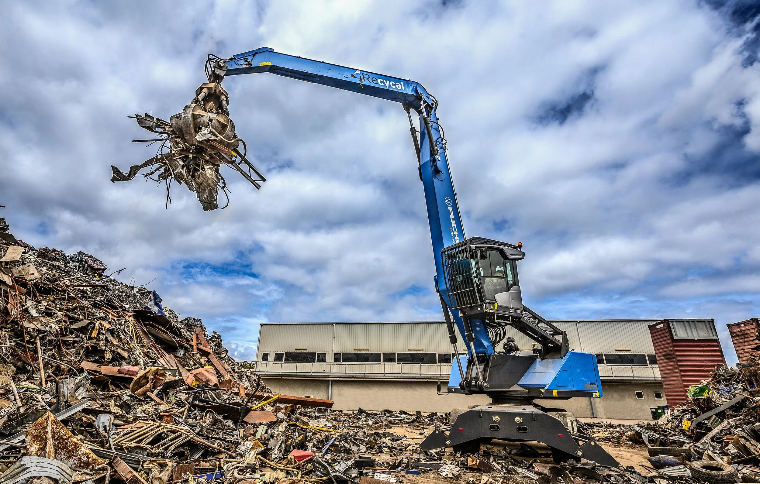 A large blue recycling excavator with a claw attachment lifting scrap metal at a recycling yard, with a building and cloudy sky in the background.