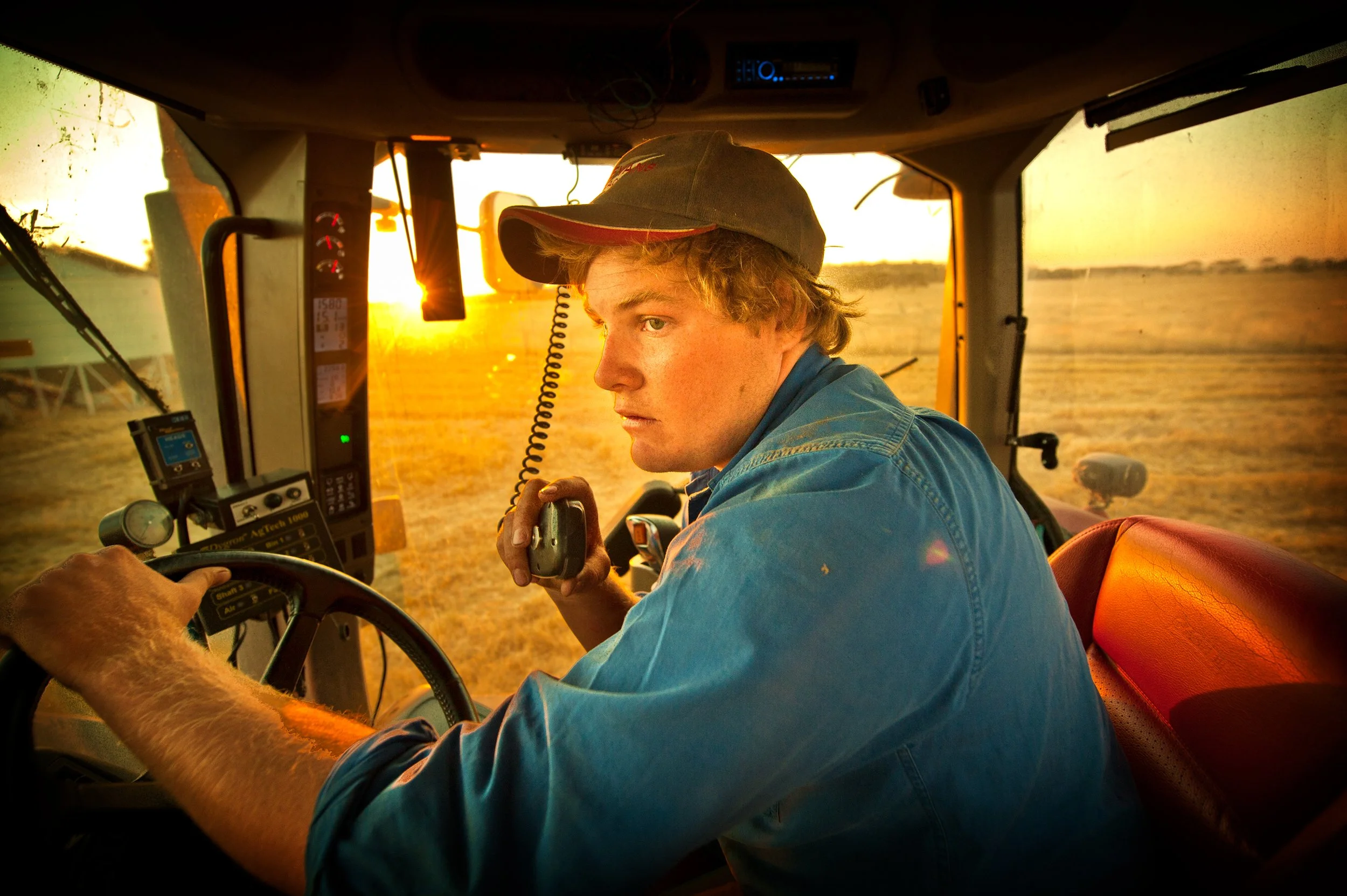 Young male farm worker driving a combine harvester or tractor through a field at sunset.