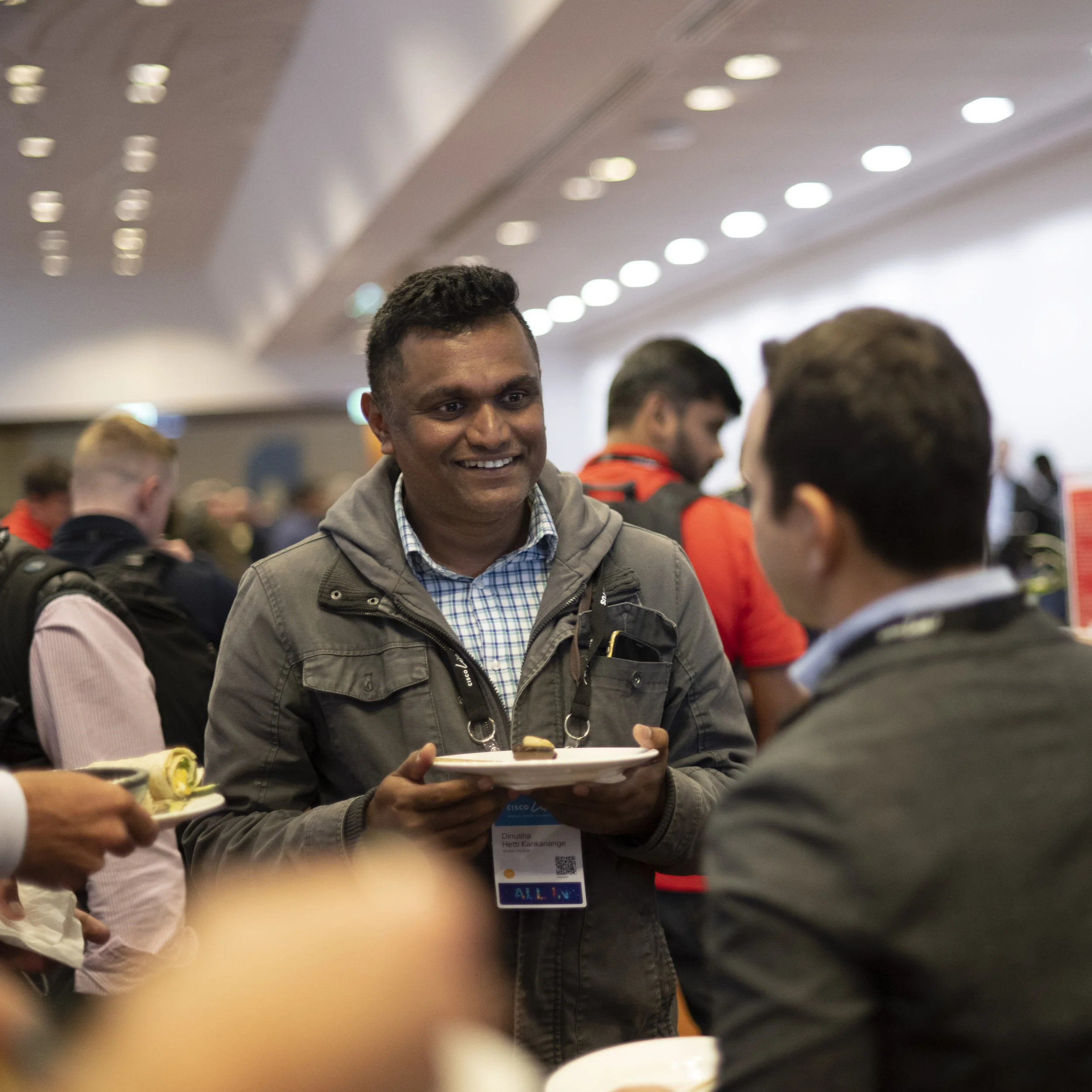 A man smiling and holding a plate with food at a conference or event, engaged in conversation with another person.