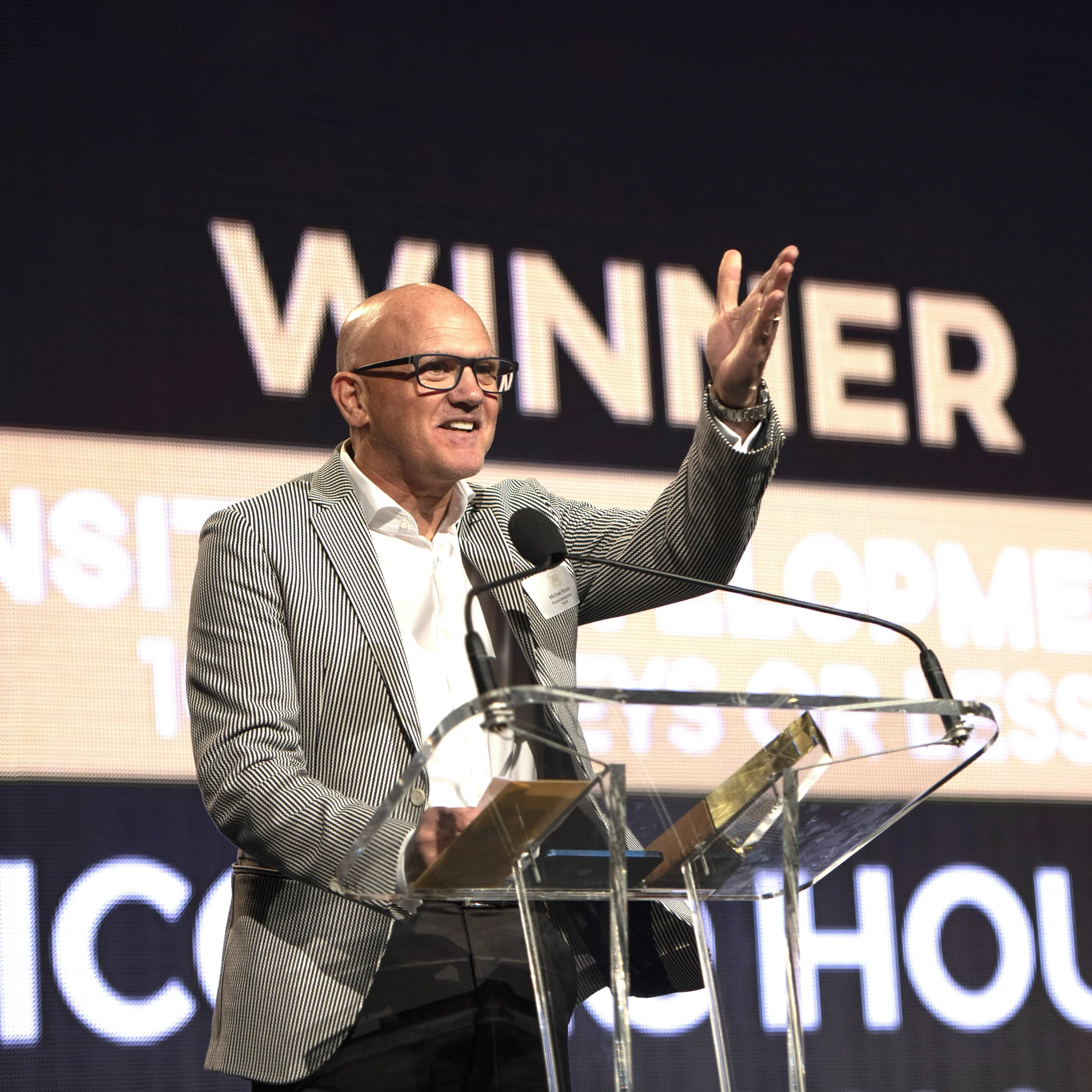 A man in a striped blazer and glasses speaking at a podium during an award ceremony, with a large screen behind him displaying the word 'WINNER' and other text.